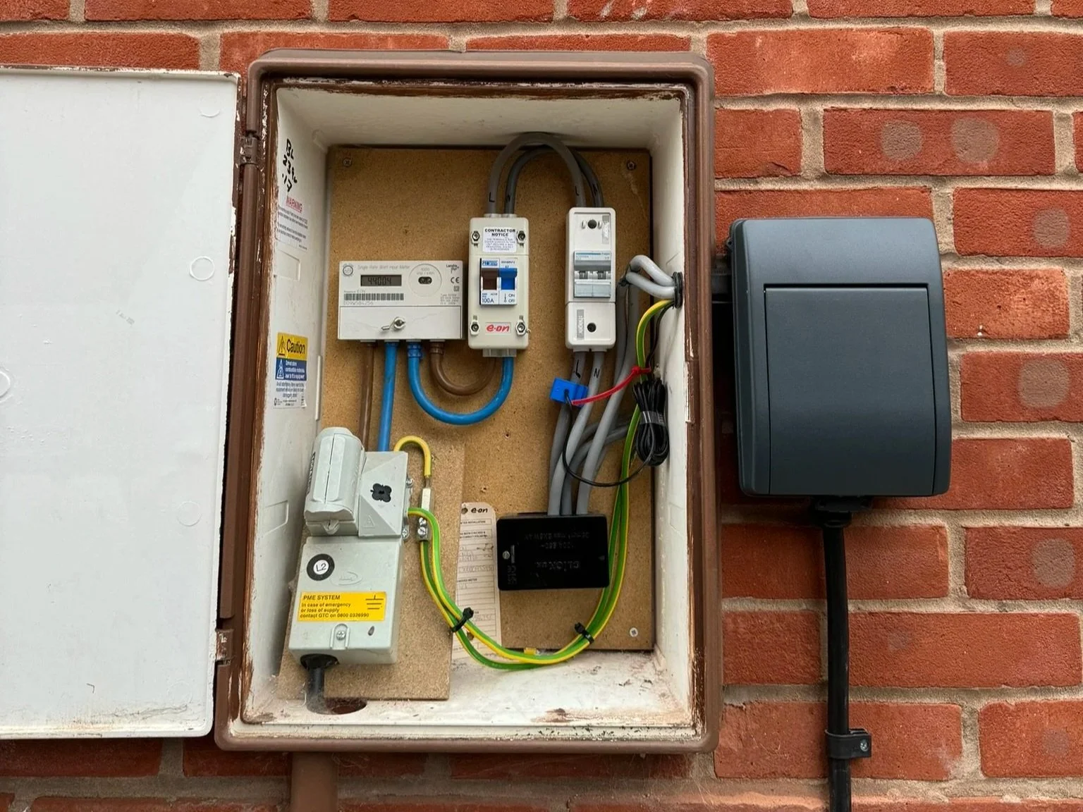 Open electrical meter box mounted on a brick wall containing an electric meter, circuit breakers, wiring, and a relay, with an external black weatherproof enclosure connected by a conduit.