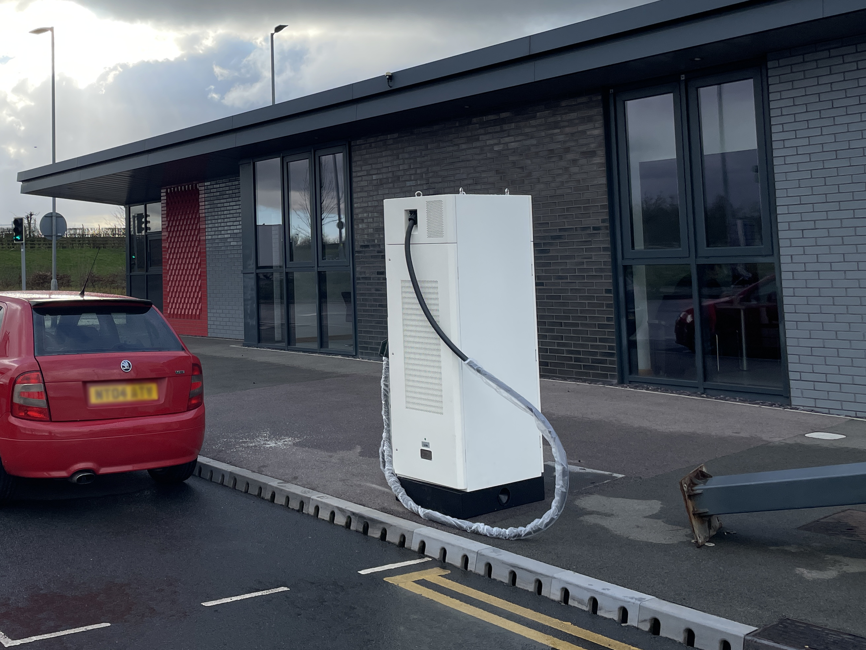 An electric vehicle charging station outside a modern commercial building with glass windows and brick walls, next to a red car, on a paved parking lot.