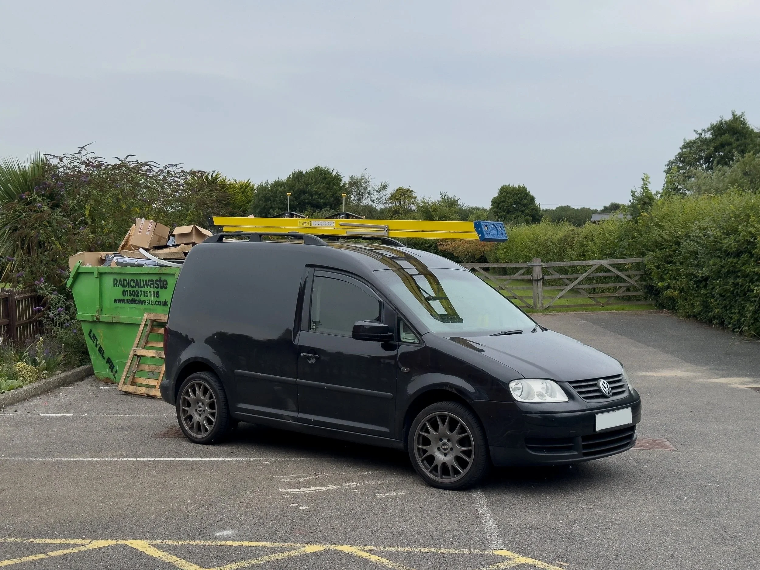 Black Volkswagen Caddy parked in a parking lot next to a green waste bin filled with trash and cardboard, with a yellow ladder on top of the van, and signaling a rural background with trees and a wooden fence.