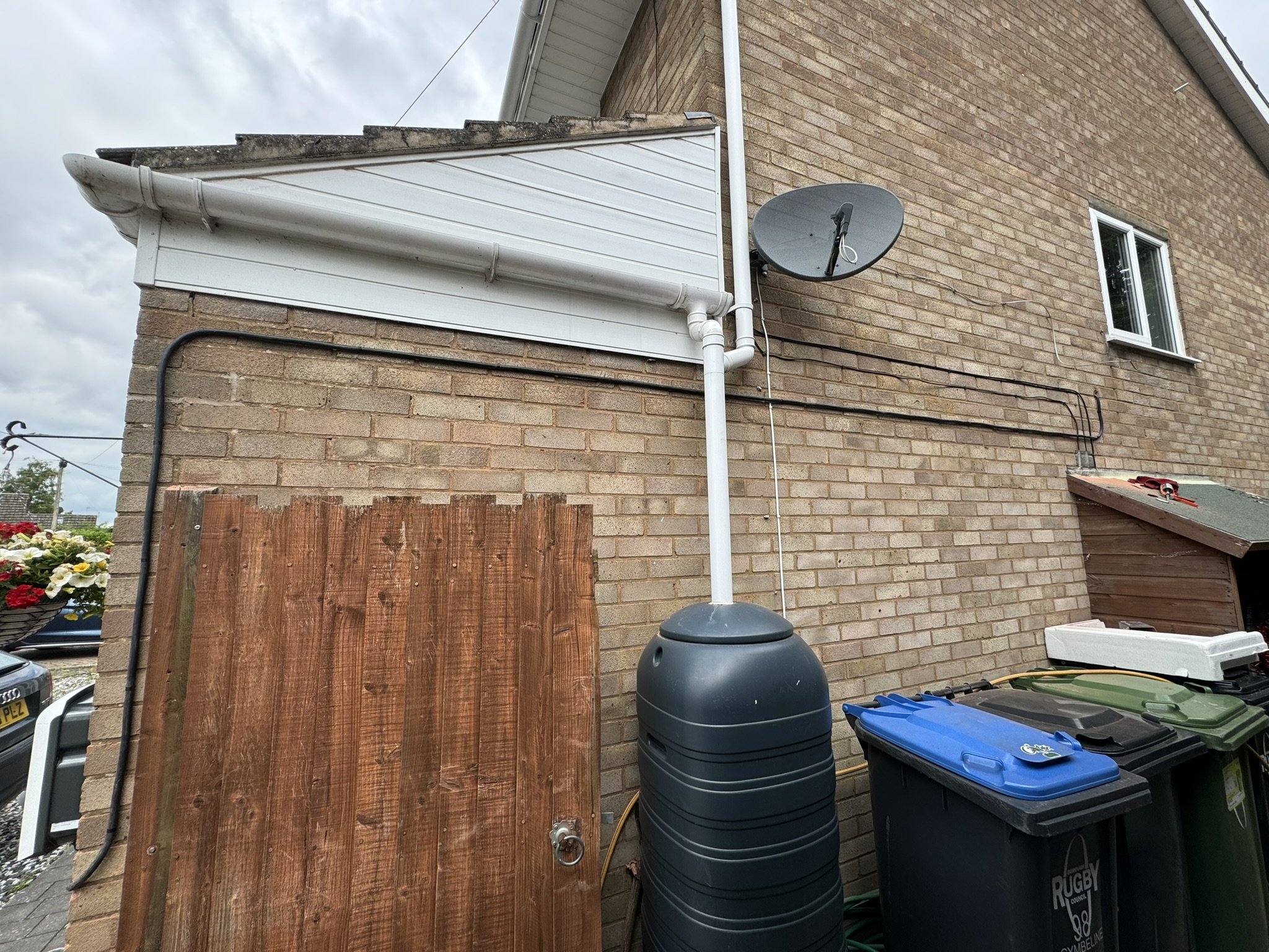 Side of a brick house with a satellite dish, white gutter, black electrical wiring, a rainwater collection tank, a green and a black garbage bin, and a wooden gate, under a cloudy sky.