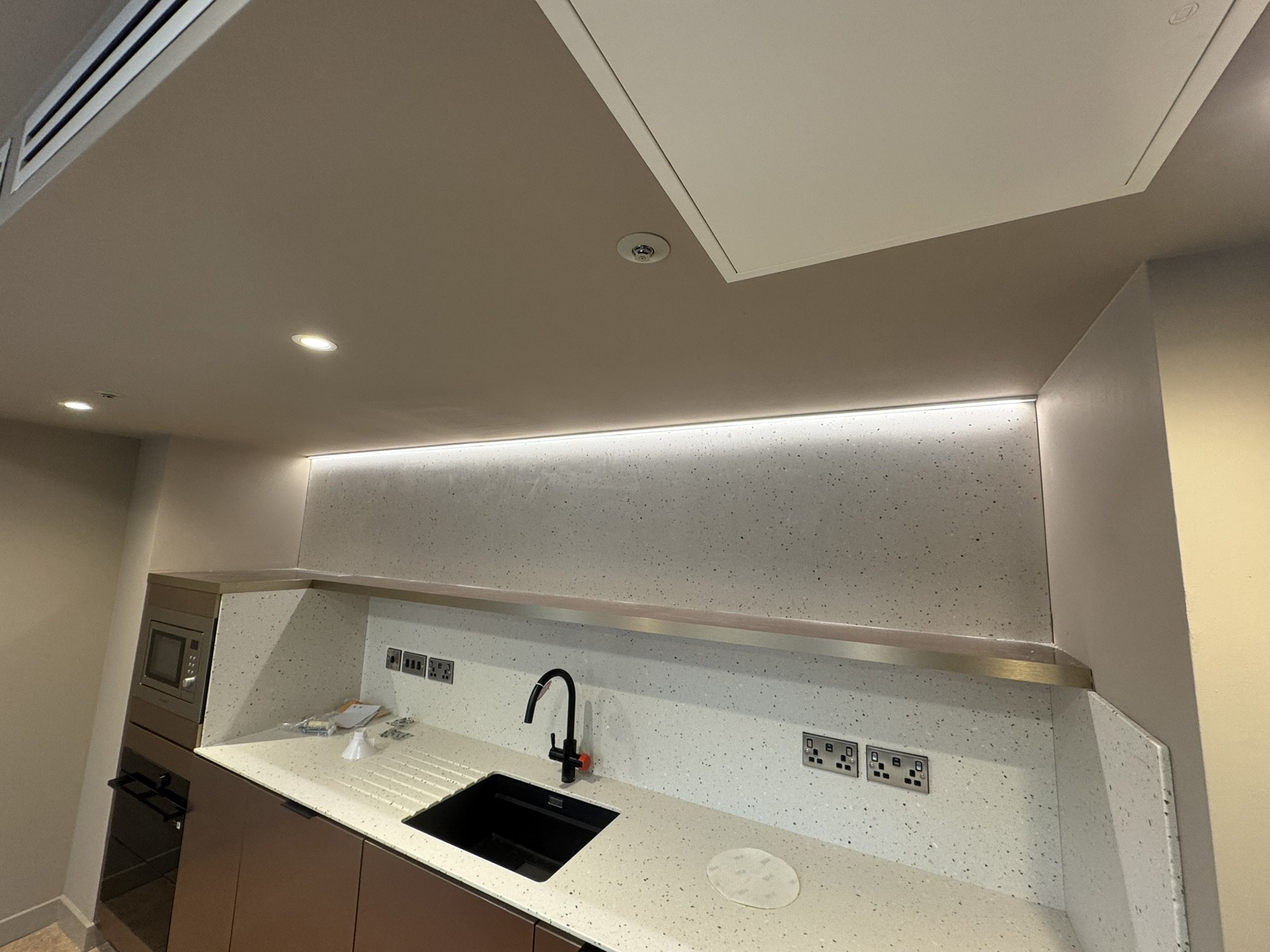 Modern kitchen with a black sink, a black faucet, and multiple electrical outlets on a speckled white countertop. The wall backsplash matches the countertop design, with a stainless steel trim. Overhead are a beige ceiling with recessed lighting and 