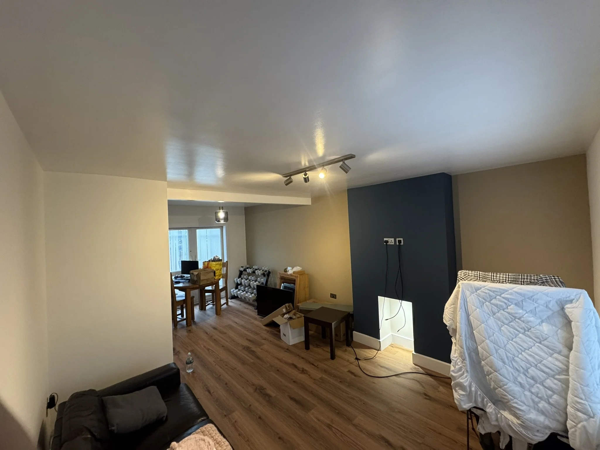 A living room with a black sofa, wooden flooring, a dining table with chairs, a window, and various furniture items and boxes.
