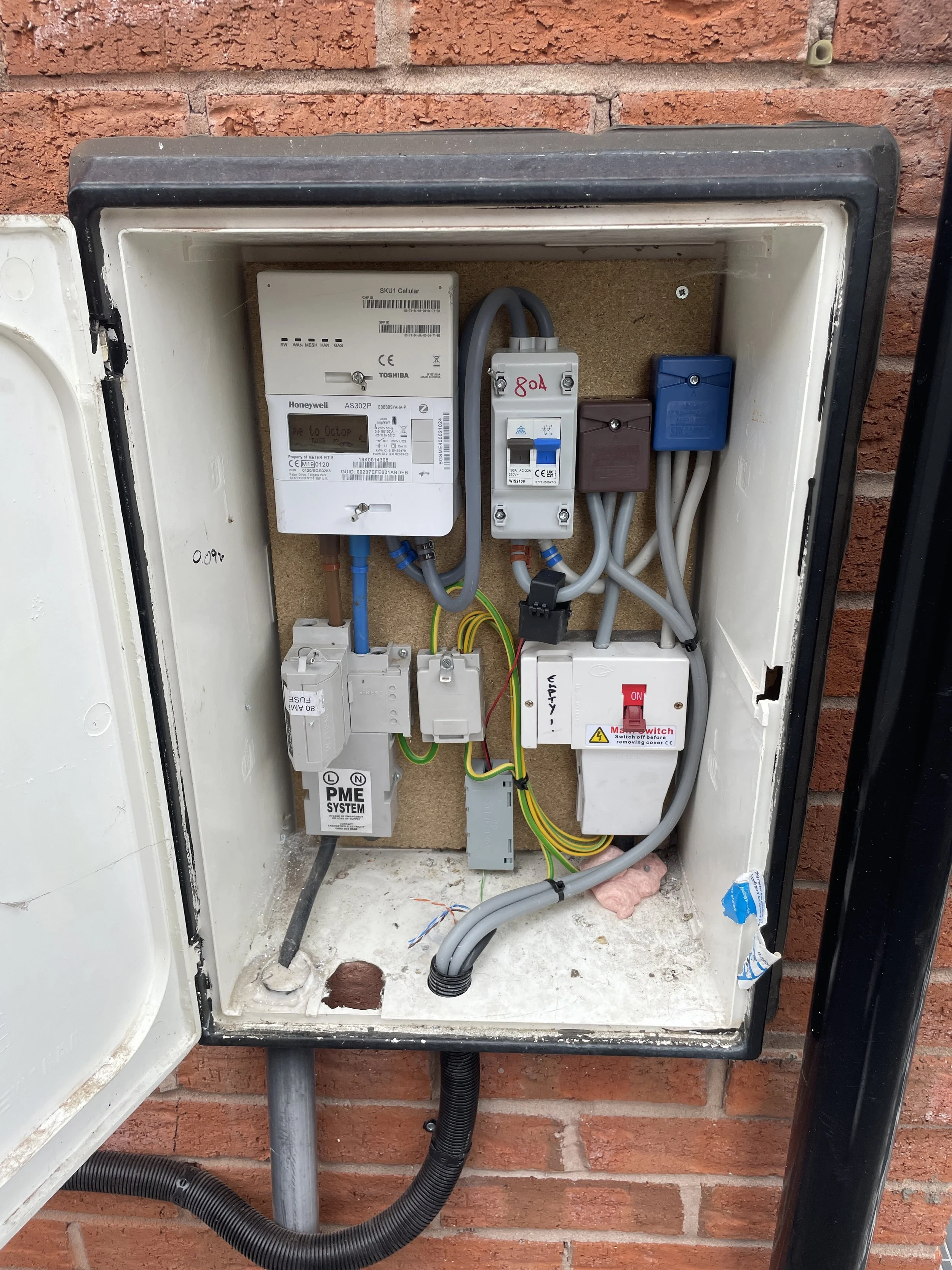 Electrical meter box with various meters, switches, and wires mounted inside against brick wall.