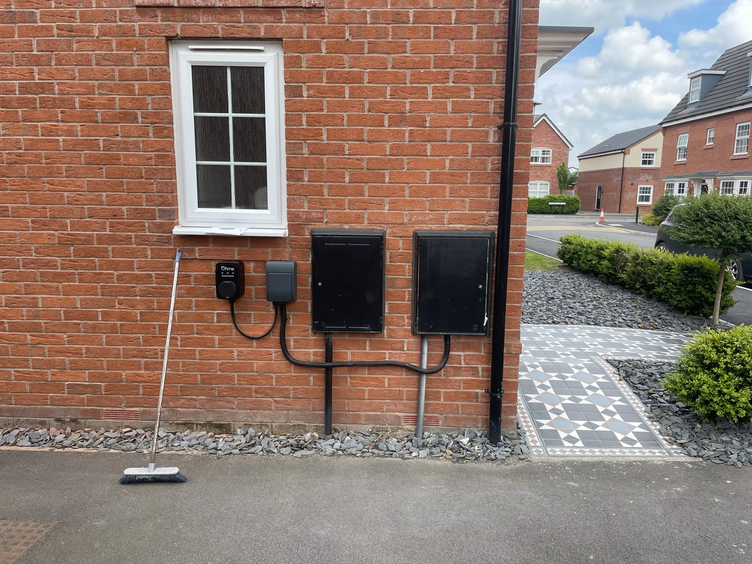 Exterior view of a brick house wall with a window, a broom leaning against the wall, and utility boxes and wires attached to the wall. A sidewalk with a patterned tile pathway and nearby greenery is visible.