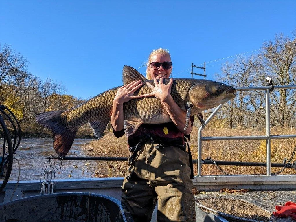 A woman wearing sunglasses and outdoor clothing on a boat, holding a large fish she caught during a fishing trip, with trees and a clear blue sky in the background.