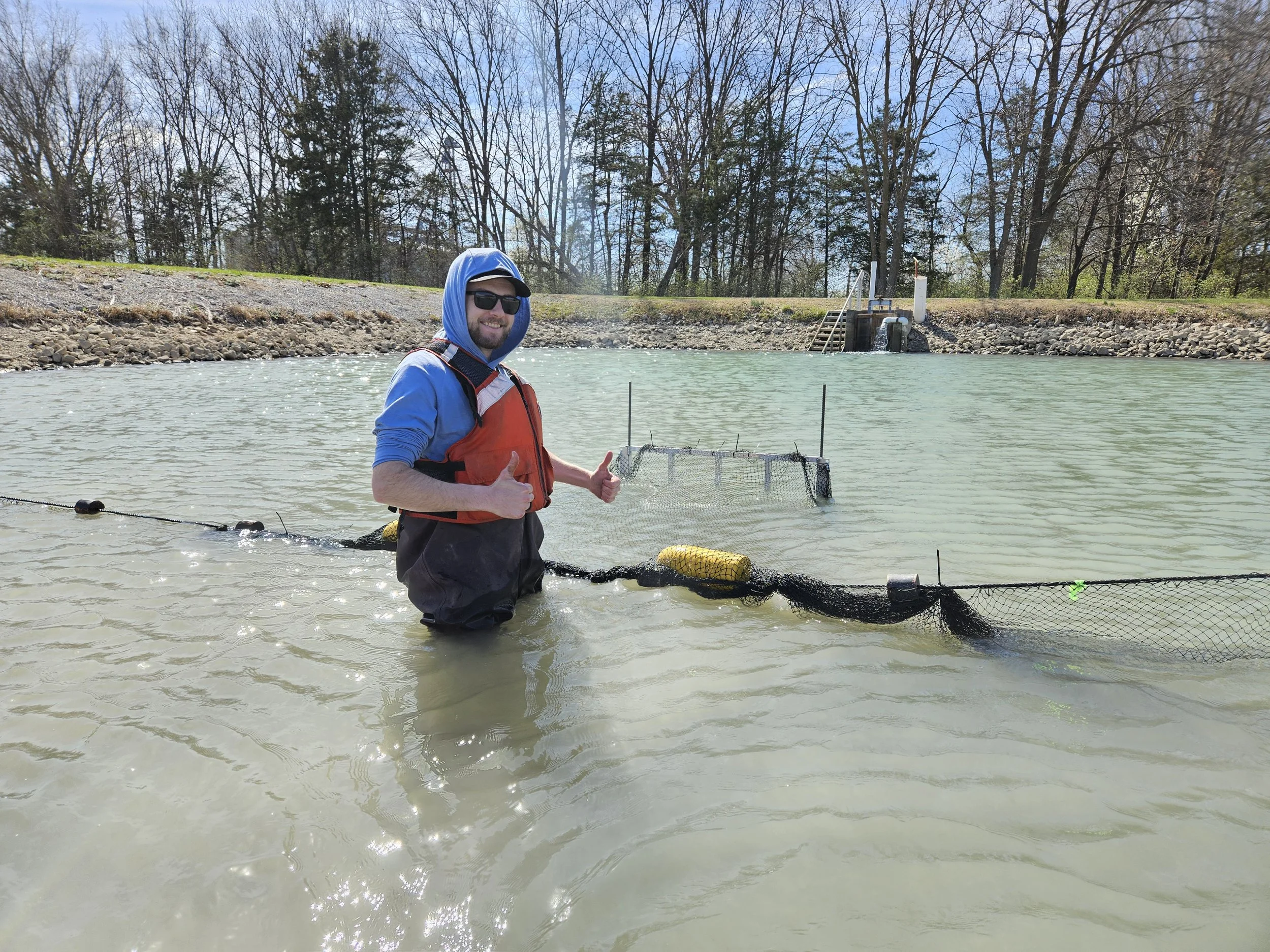 A man standing in a river, giving a thumbs-up, wearing sunglasses, a hooded blue jacket, a life vest, and waders. He is near a fish trap or net, with a small dam and a waterfall structure in the background under a partly cloudy sky.