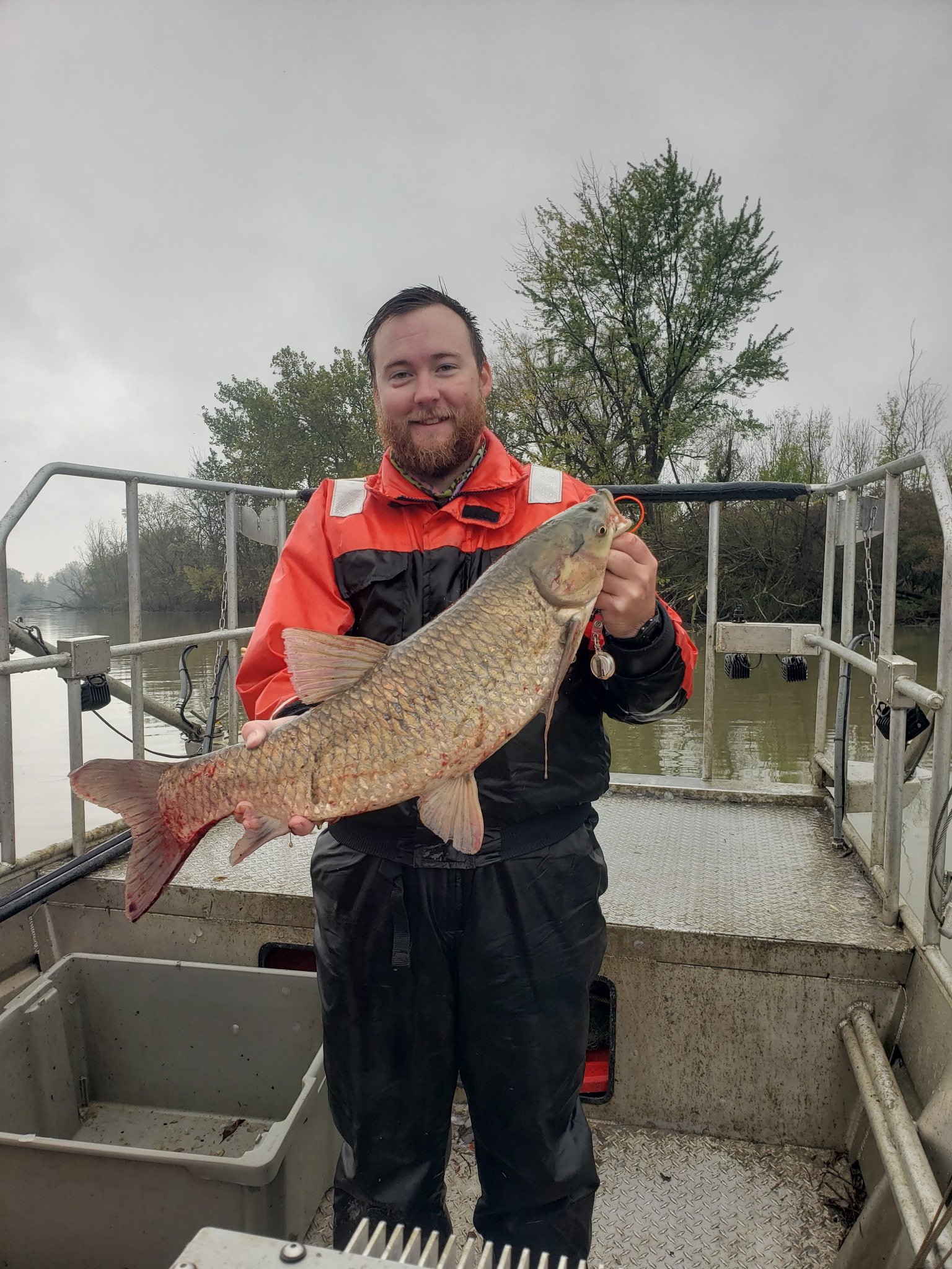 Man in a red and black jacket smiling and holding a large fish on a boat.