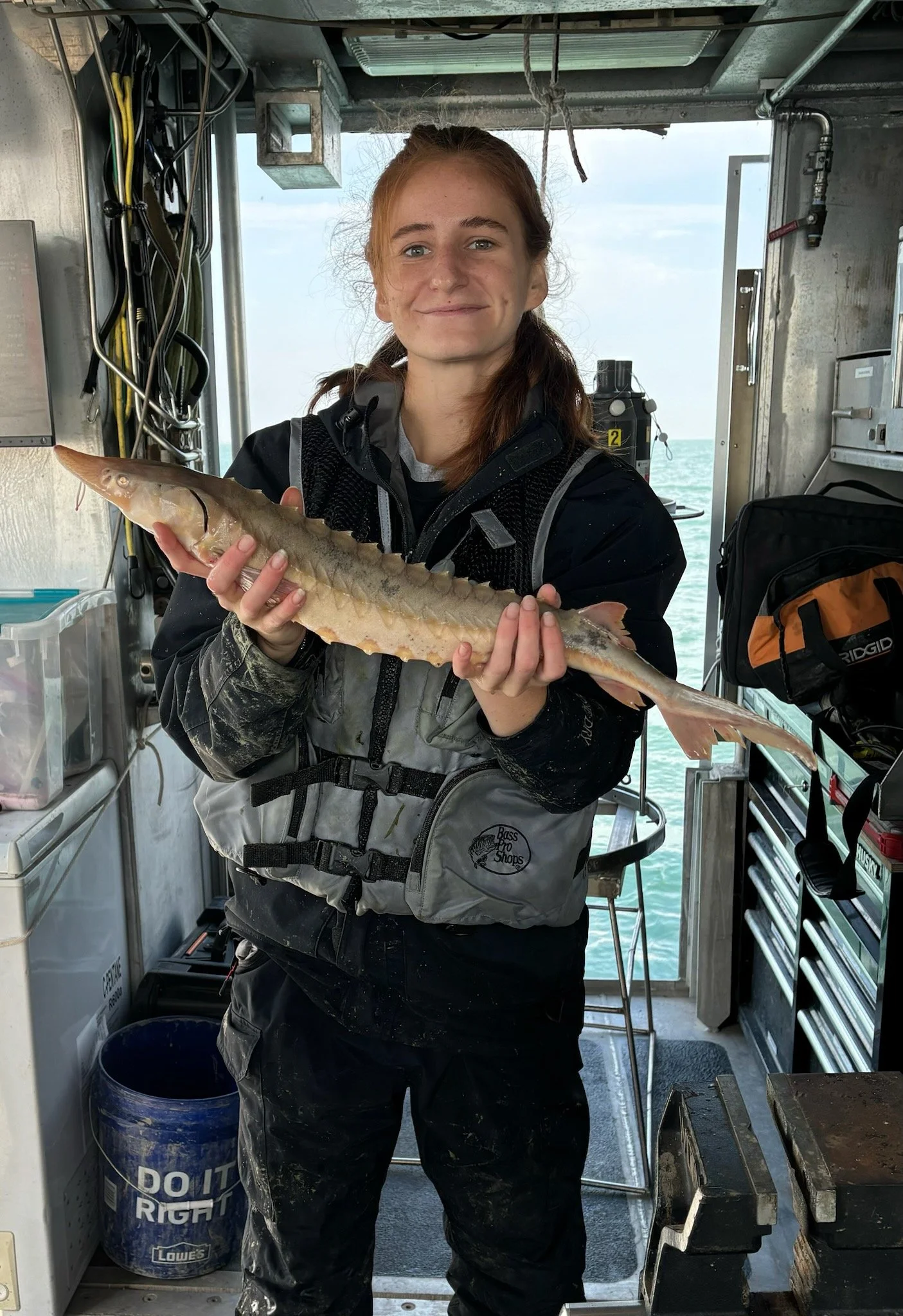 A young woman with red hair smiling and holding a large fish inside a boat or fishing vessel, with water and sky visible in the background.