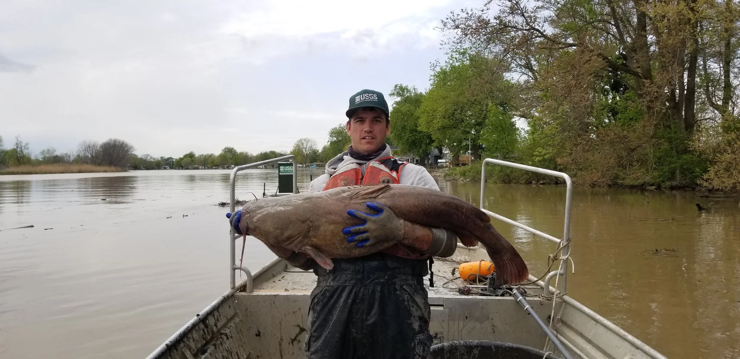 Young man holding a large fish on a boat with water and trees in the background, wearing a USGS hat and gloves.