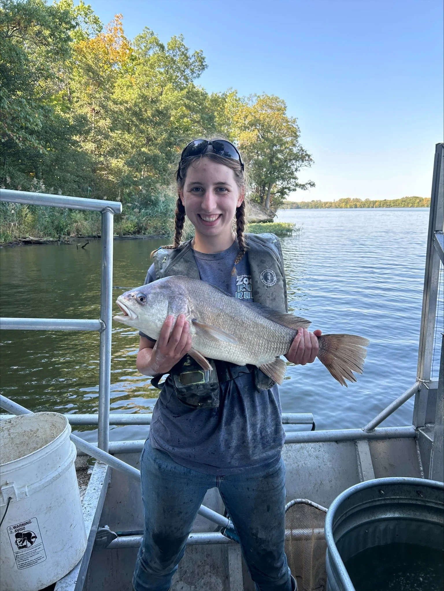 A young girl with braids and sunglasses on her head holding a large fish on a boat by a river with trees in the background.