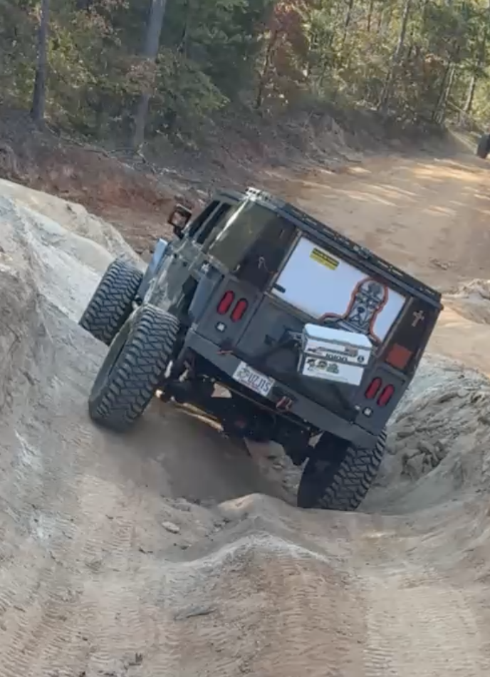 A black off-road Jeep driving through a rugged dirt trail with deep ruts and uneven terrain surrounded by trees.