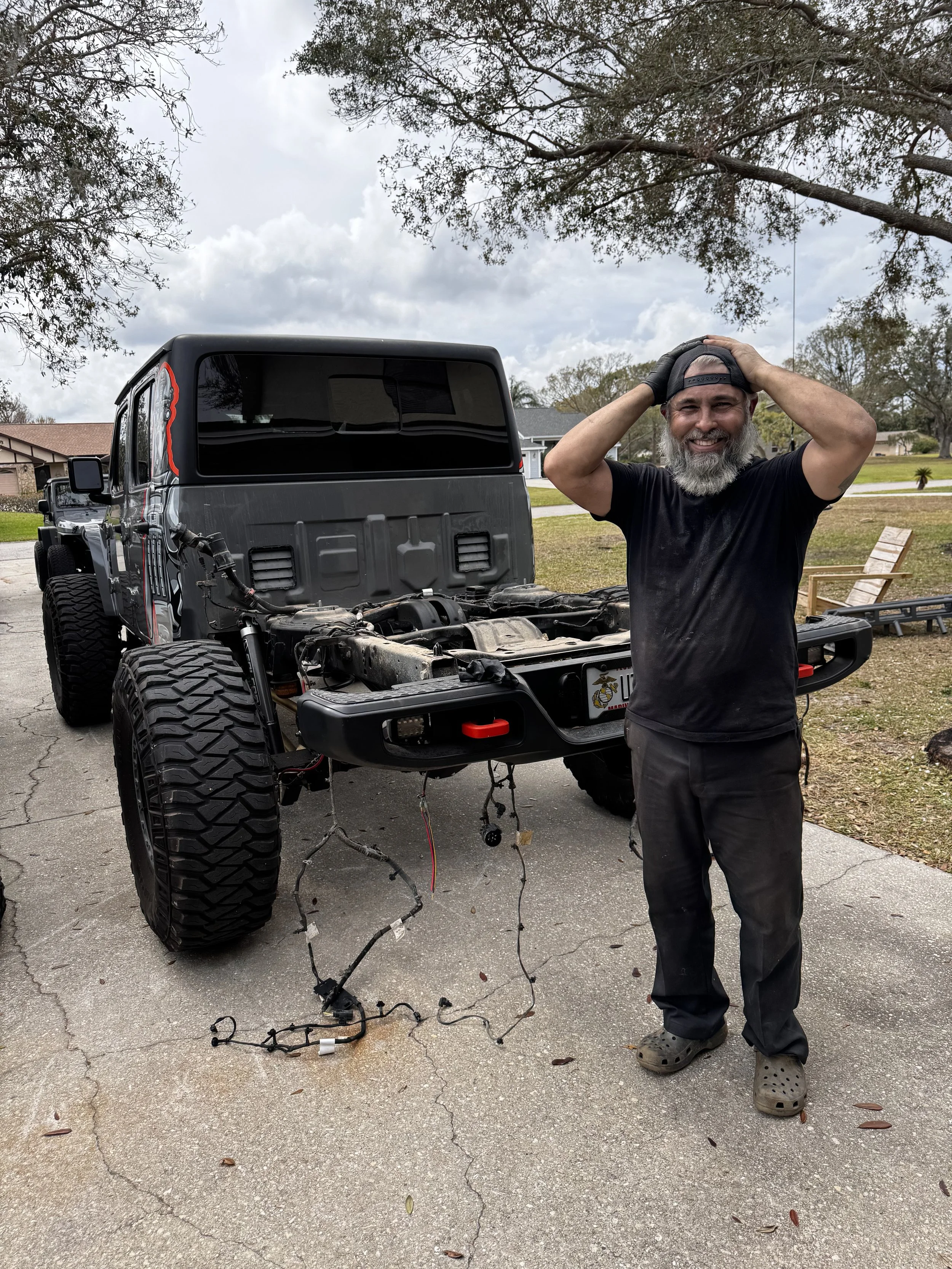 A man with a beard and black shirt standing outdoors next to a black off-road vehicle with missing engine parts. He is smiling and holding his cap with both hands. The vehicle appears to be under repair or modification, with exposed wiring hanging from the front.