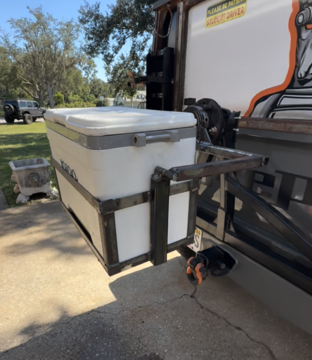 A metal frame mounted on a vehicle holding a white cooler and an ice chest, with a sticker that says 'Please be patient, student driver' and an illustration of a penguin in the background.
