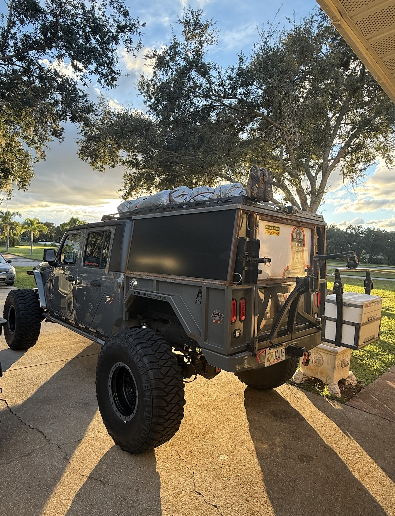 A gray off-road vehicle with large tires parked on a driveway, loaded with gear on top, a large storage box on the back, and surrounded by trees, during sunset.
