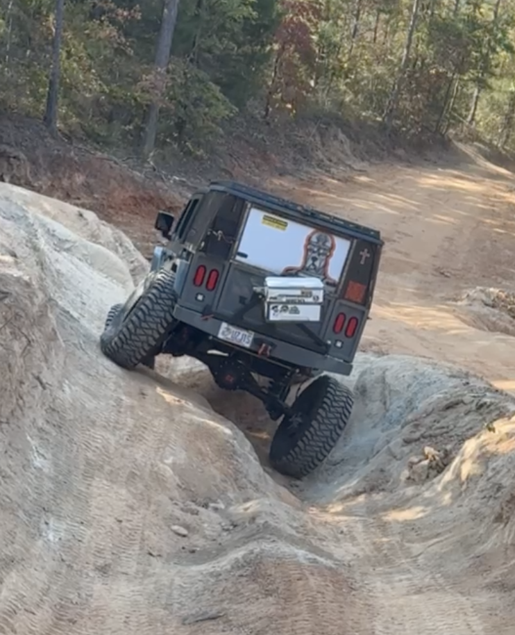 A black off-road vehicle navigating a rugged dirt trail with deep tire tracks and uneven terrain, surrounded by trees and foliage.