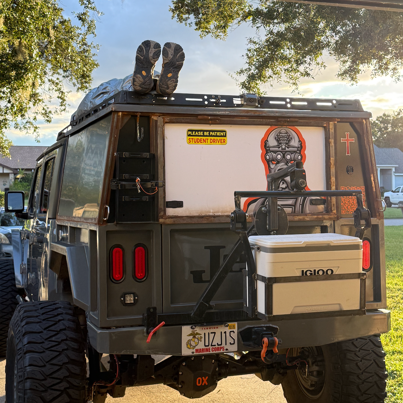 The back of a custom off-road vehicle with a person lying on top, visible only from the legs and boots. The vehicle has various stickers and equipment, including a sign that says 'Please Be Patient Student Driver,' a cartoon-style robot graphic, and a cooler mounted on the rear.