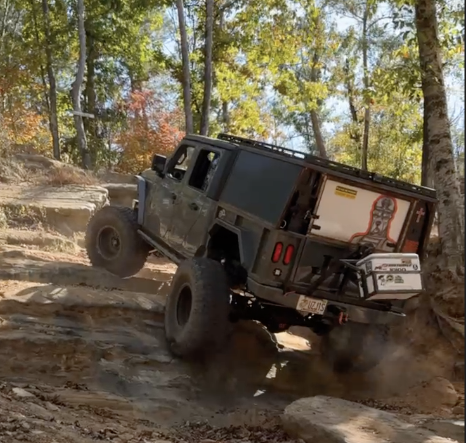 A black off-road vehicle climbing a rocky and muddy trail in a forest with colorful autumn leaves and sunlight filtering through trees.