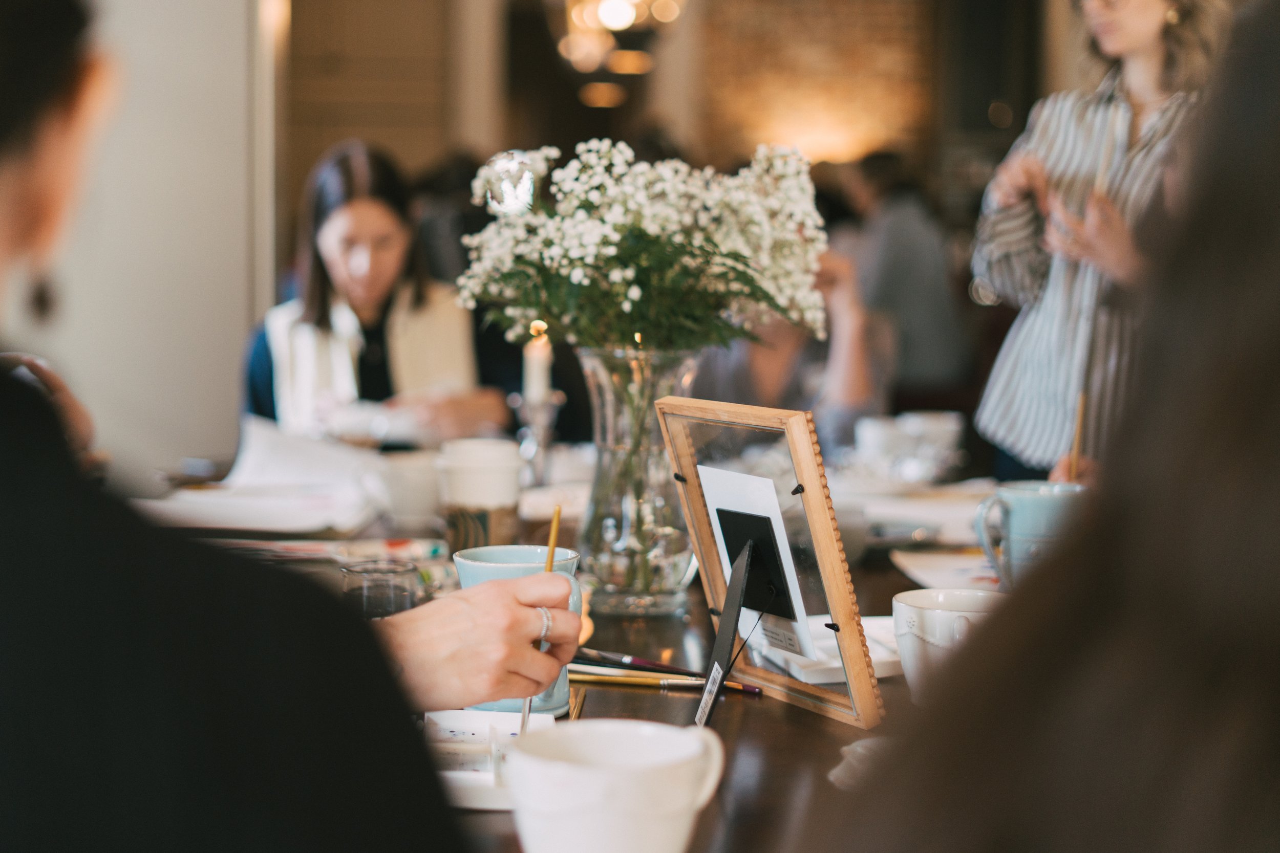 People gathering around a table with a large vase of white flowers, cups, and framed photos, in a warmly lit room.