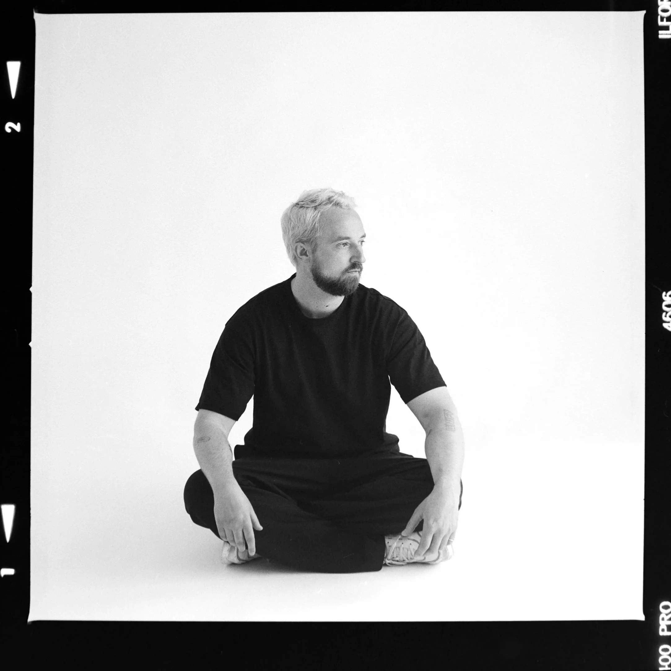 Schwarz-weiß-Fotografie von Michael Schwarz in Bayreuth schwarzen T-Shirt, sitzend und nach rechts schauend, vor einem weißen Hintergrund. Ein Teil seines Körpers ist sichtbar.