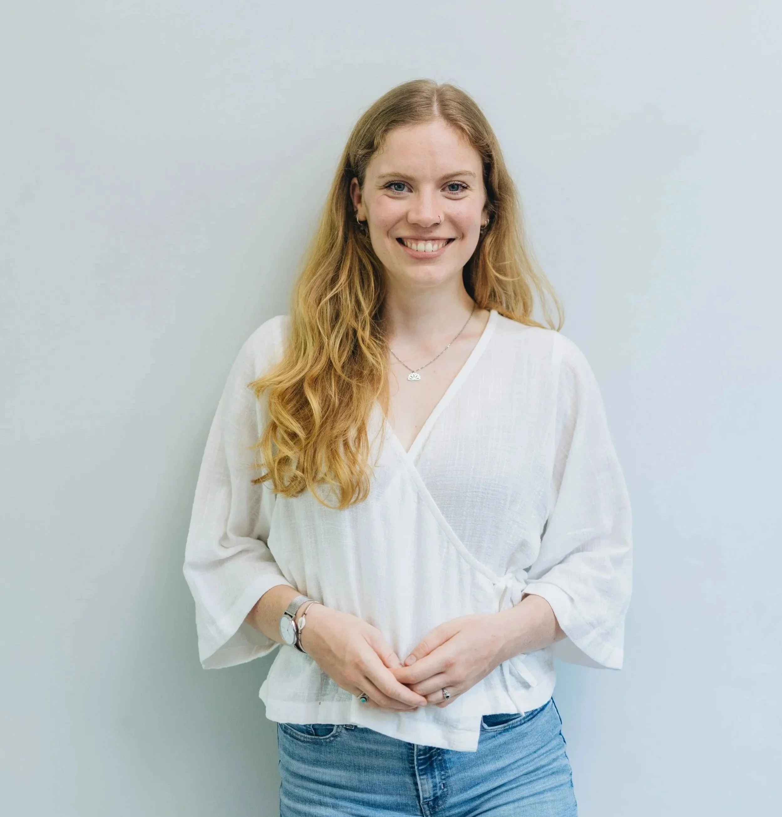 A young woman with long, wavy red hair, smiling, wearing a white blouse and blue jeans, standing against a plain light-colored wall.