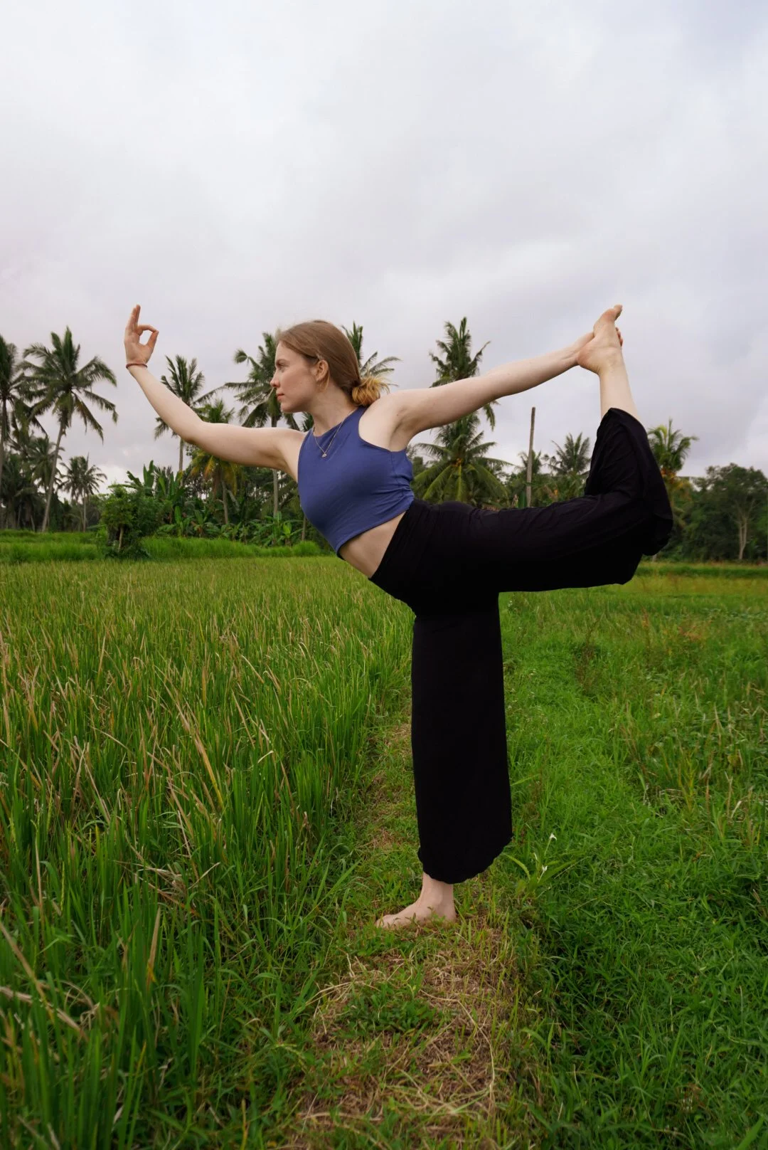 A woman practicing yoga outside in a lush green field with palm trees in the background, balancing on one leg while holding her other foot behind her, with her arms extended forward and to the side.