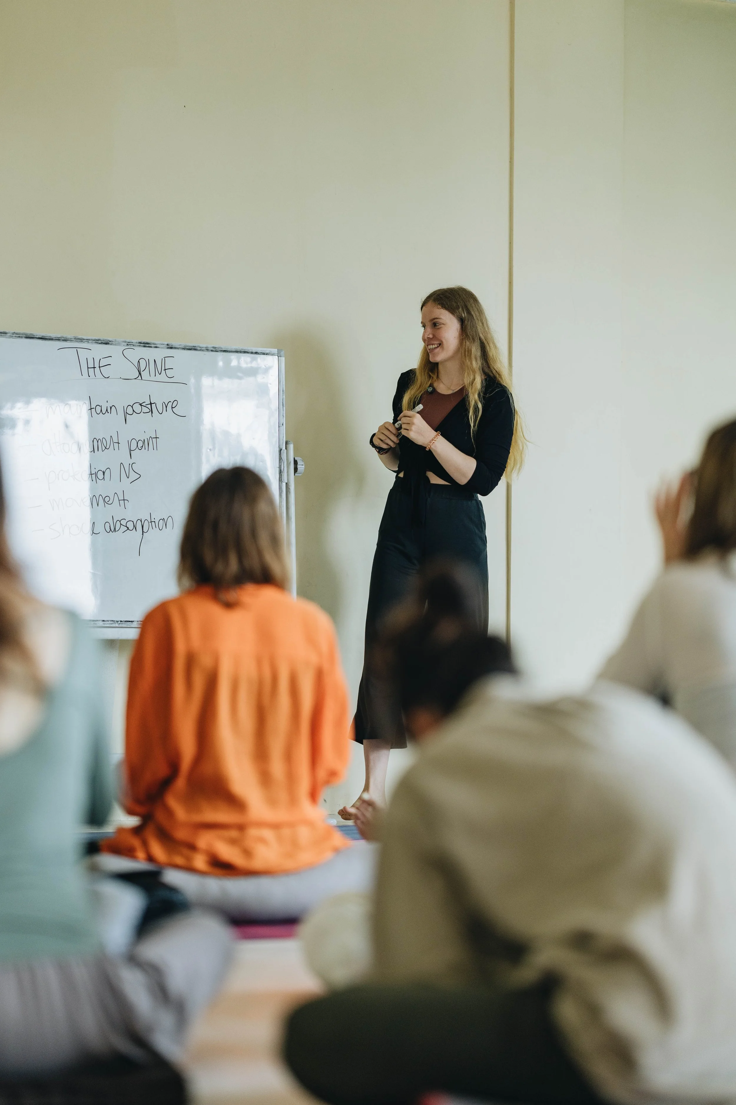 A woman stands at the front of a classroom, giving a lecture with a whiteboard behind her. Several students are seated on the floor, facing her and listening.