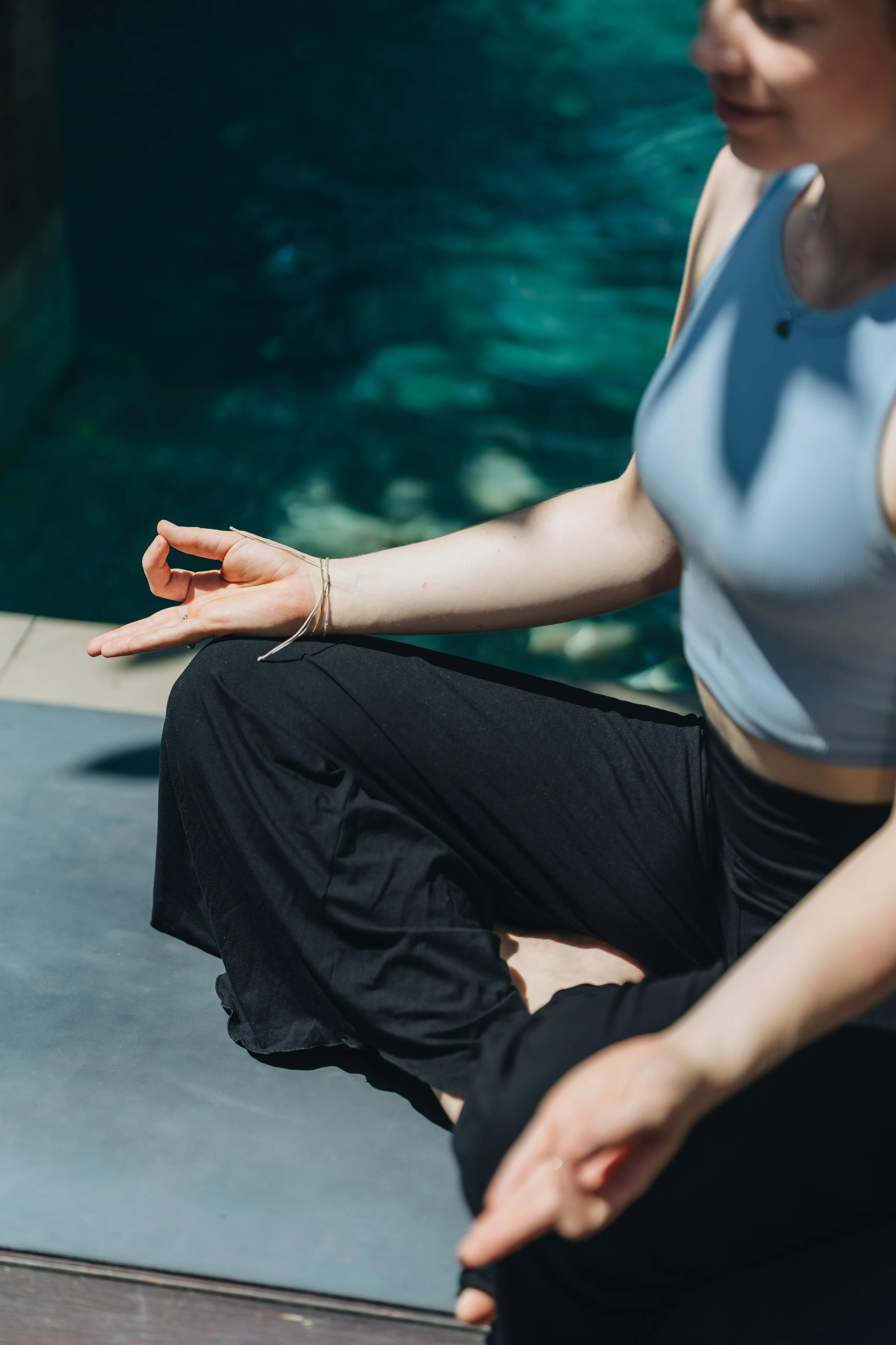 A woman practicing yoga near a pool, sitting cross-legged with her hand on her knee in a meditative pose.