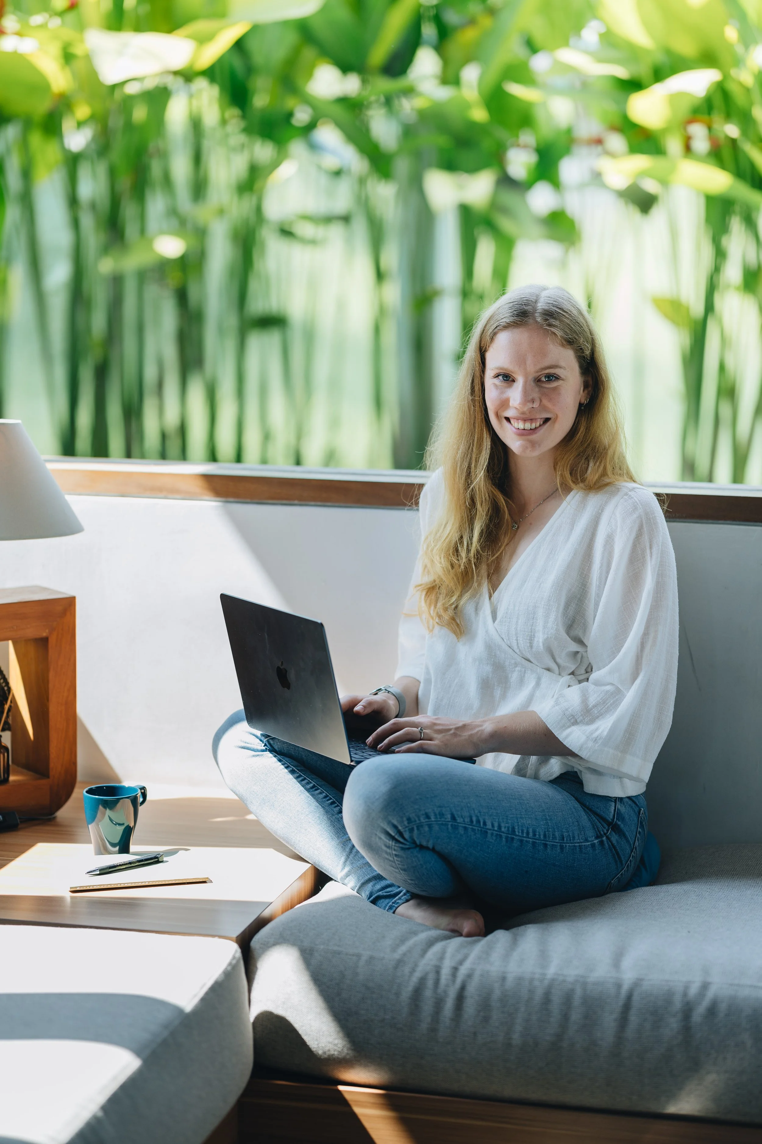 A woman sitting cross-legged on a sofa with a laptop on her lap, smiling, in a bright room with large windows showing lush green plants outside.