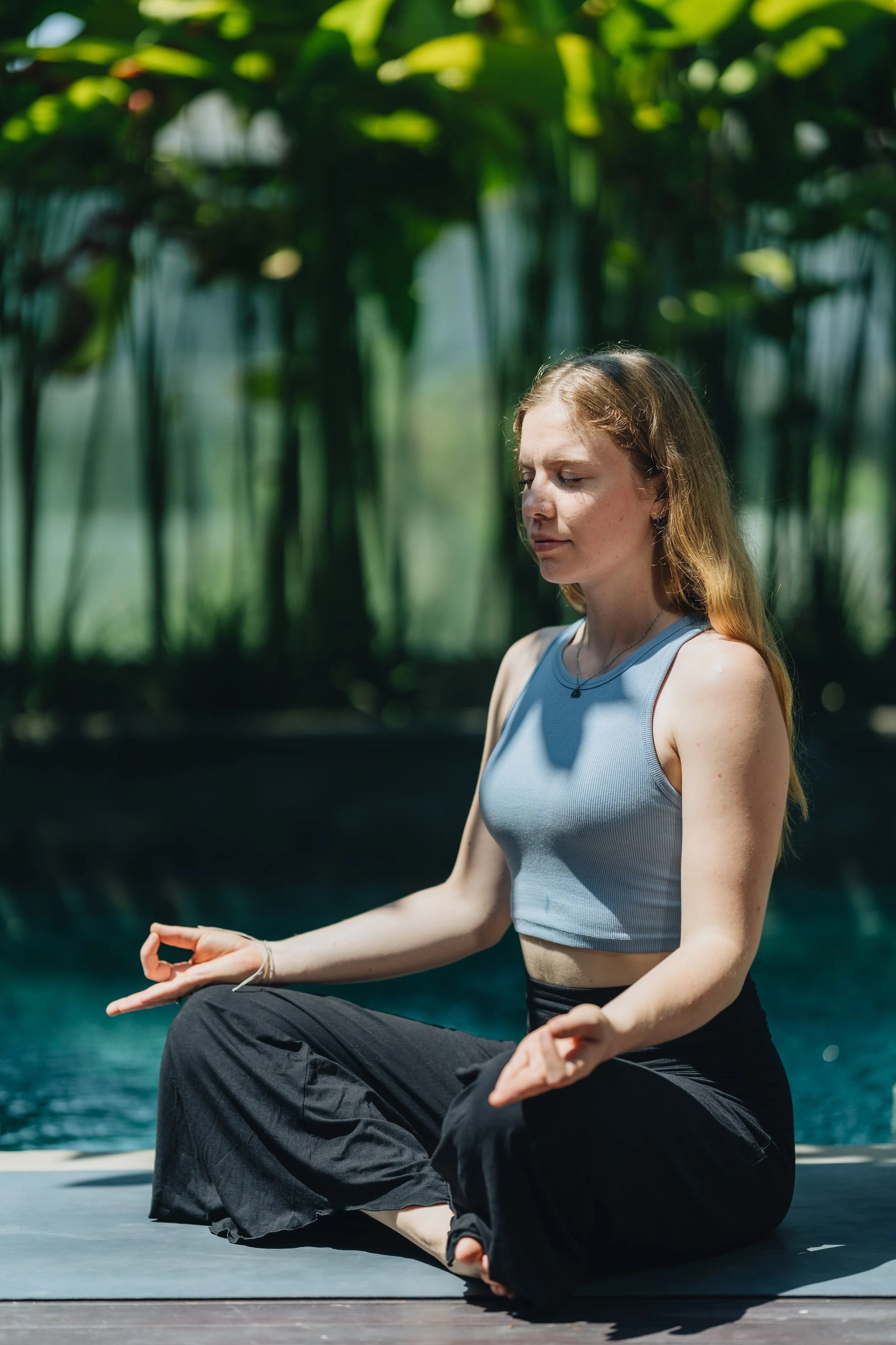 A woman practicing yoga outdoors near water with lush green plants in the background.