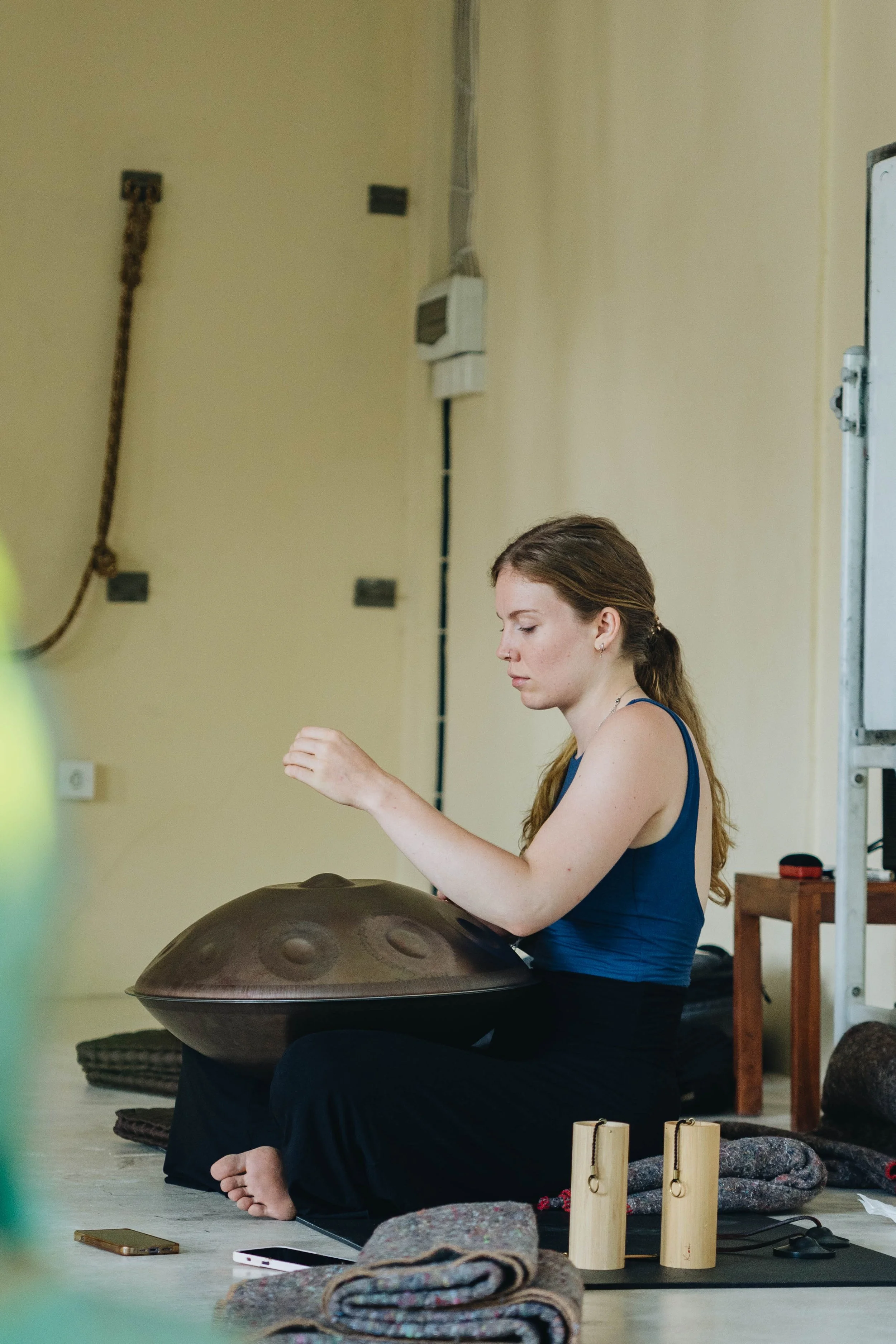 A woman sitting cross-legged on the floor, playing a handpan drum, surrounded by mats, rolled rugs, and small wooden percussion instruments.
