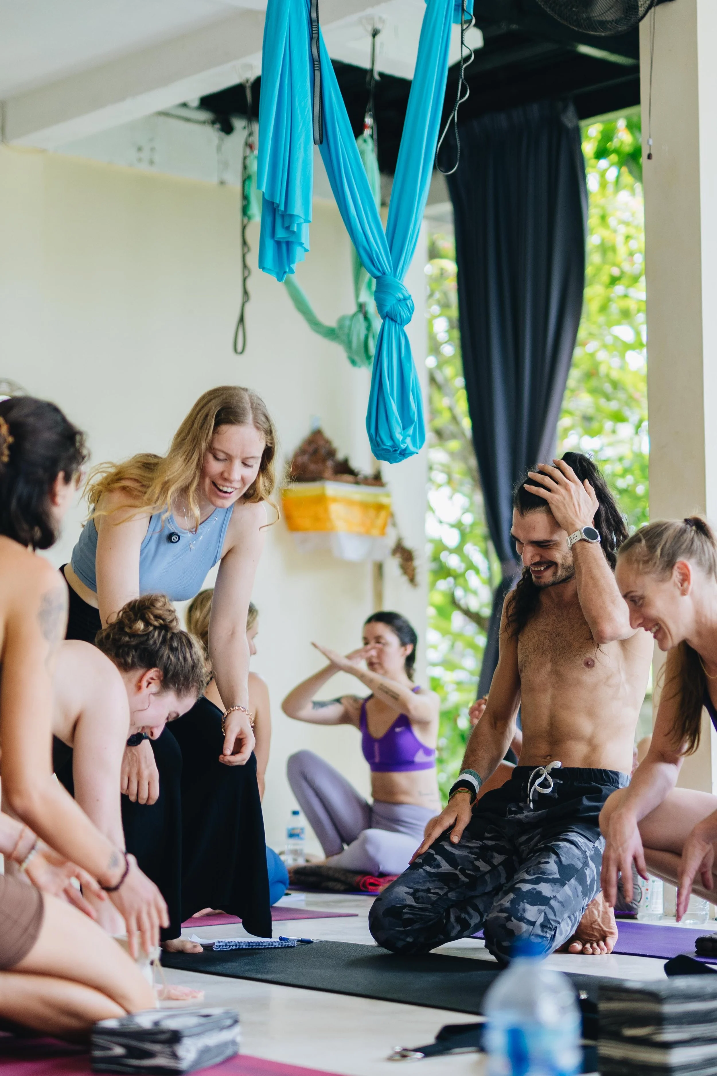 People participating in a yoga class, some are practicing poses while others laugh and interact in a bright room with blue aerial yoga hammocks hanging from the ceiling.