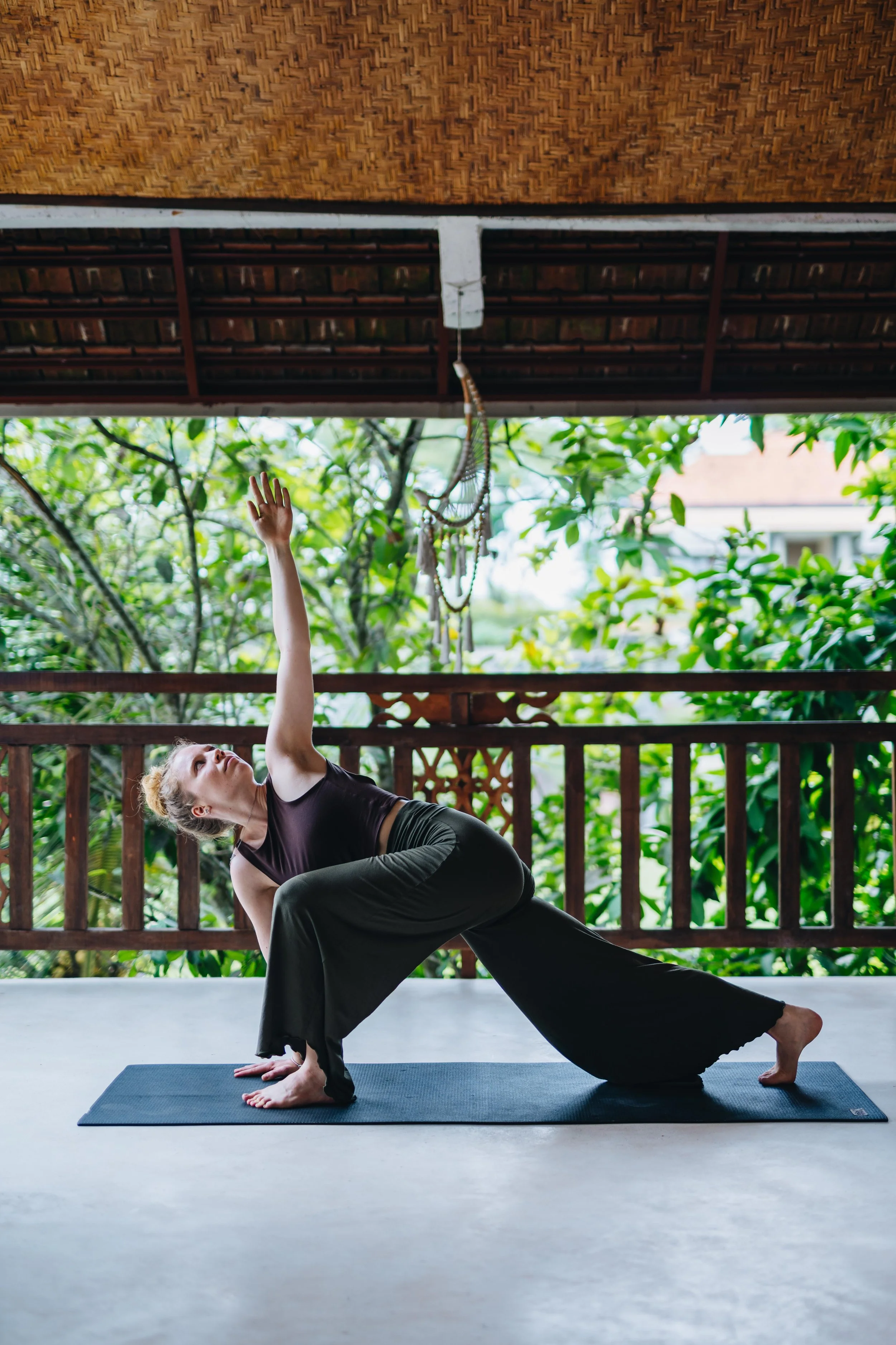 A woman practicing yoga on a mat outdoors on a porch with wooden railing, surrounded by lush green trees and foliage.