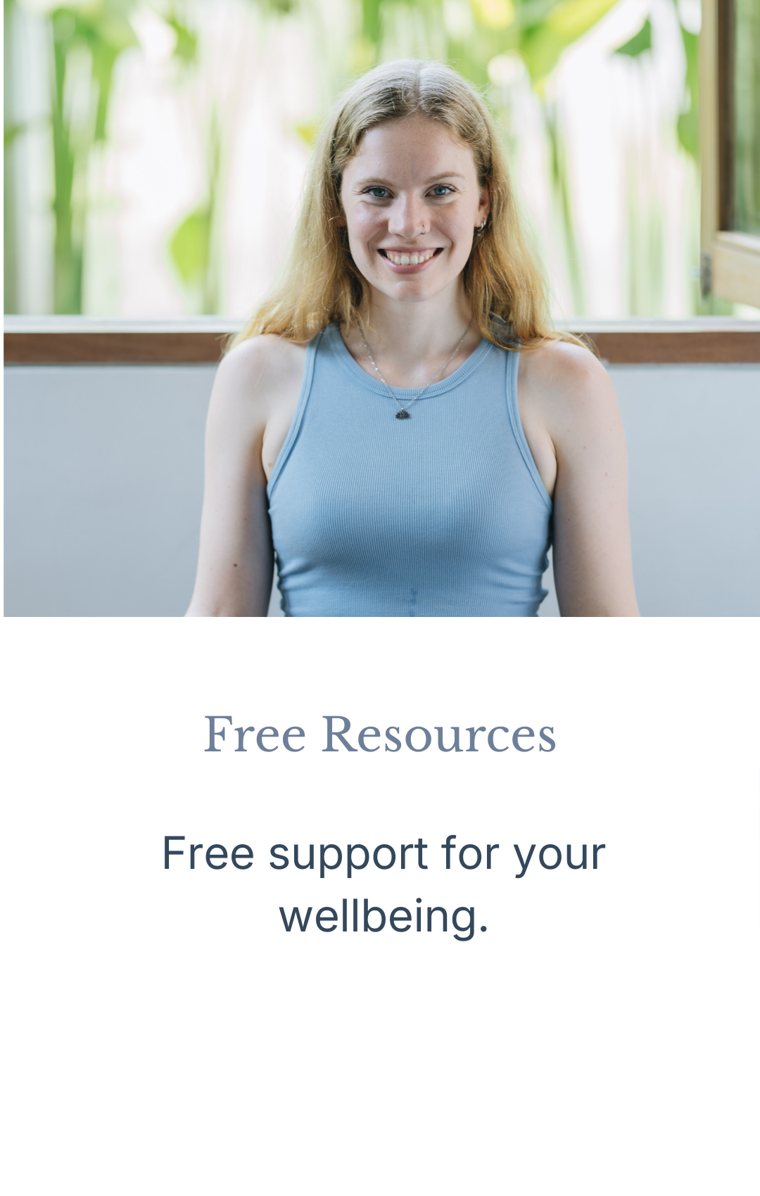 A smiling young woman with red hair wearing a blue tank top, sitting indoors with a green leafy background.