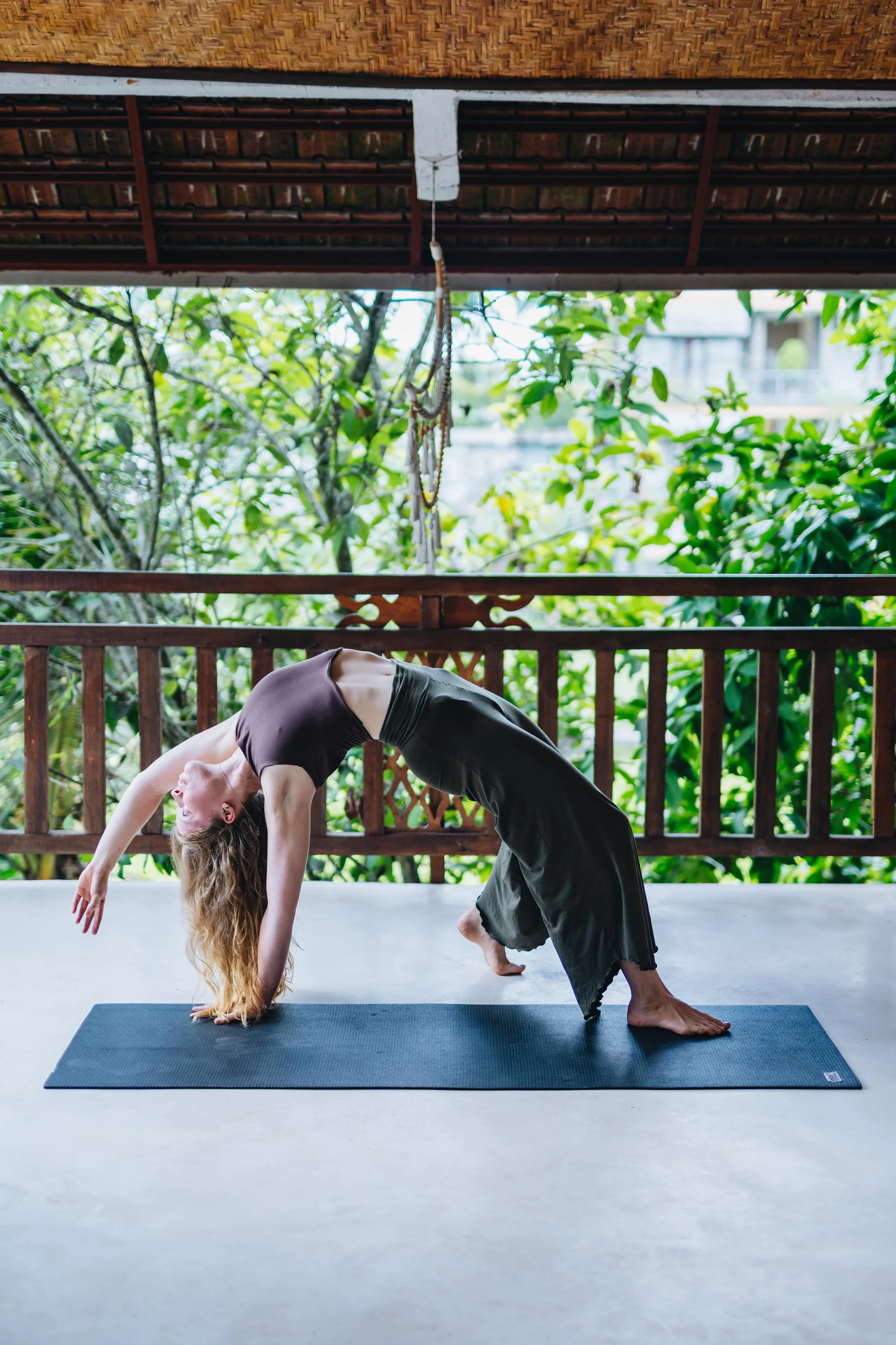 A woman practicing yoga on a black mat on a covered outdoor patio, surrounded by green trees and foliage, in a bridge pose with her hands and feet on the ground and her back arched.