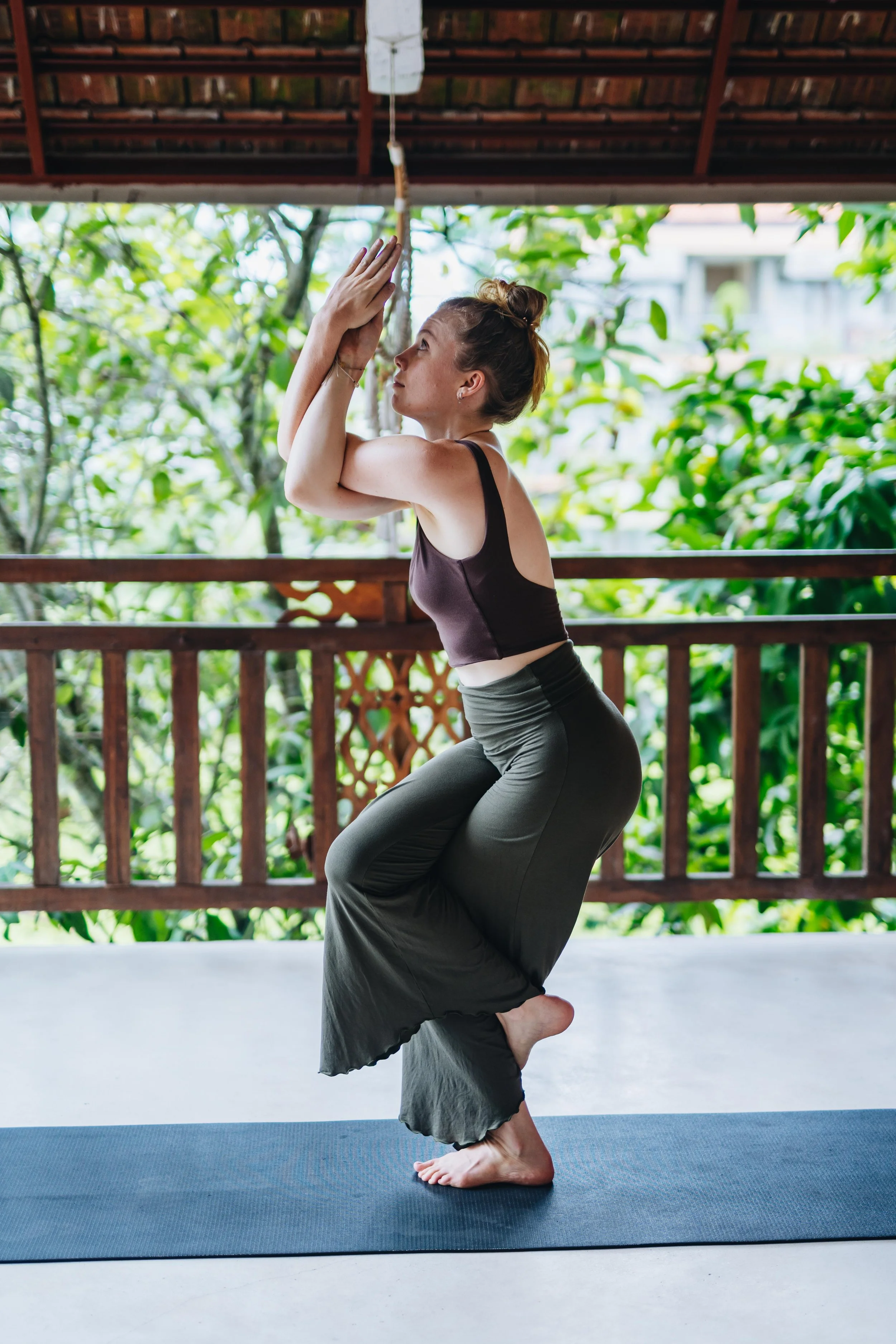 A woman practicing yoga on a black mat outdoors, standing on one foot in a tree pose, with her hands pressed together in prayer position, surrounded by green foliage and a wooden railing.