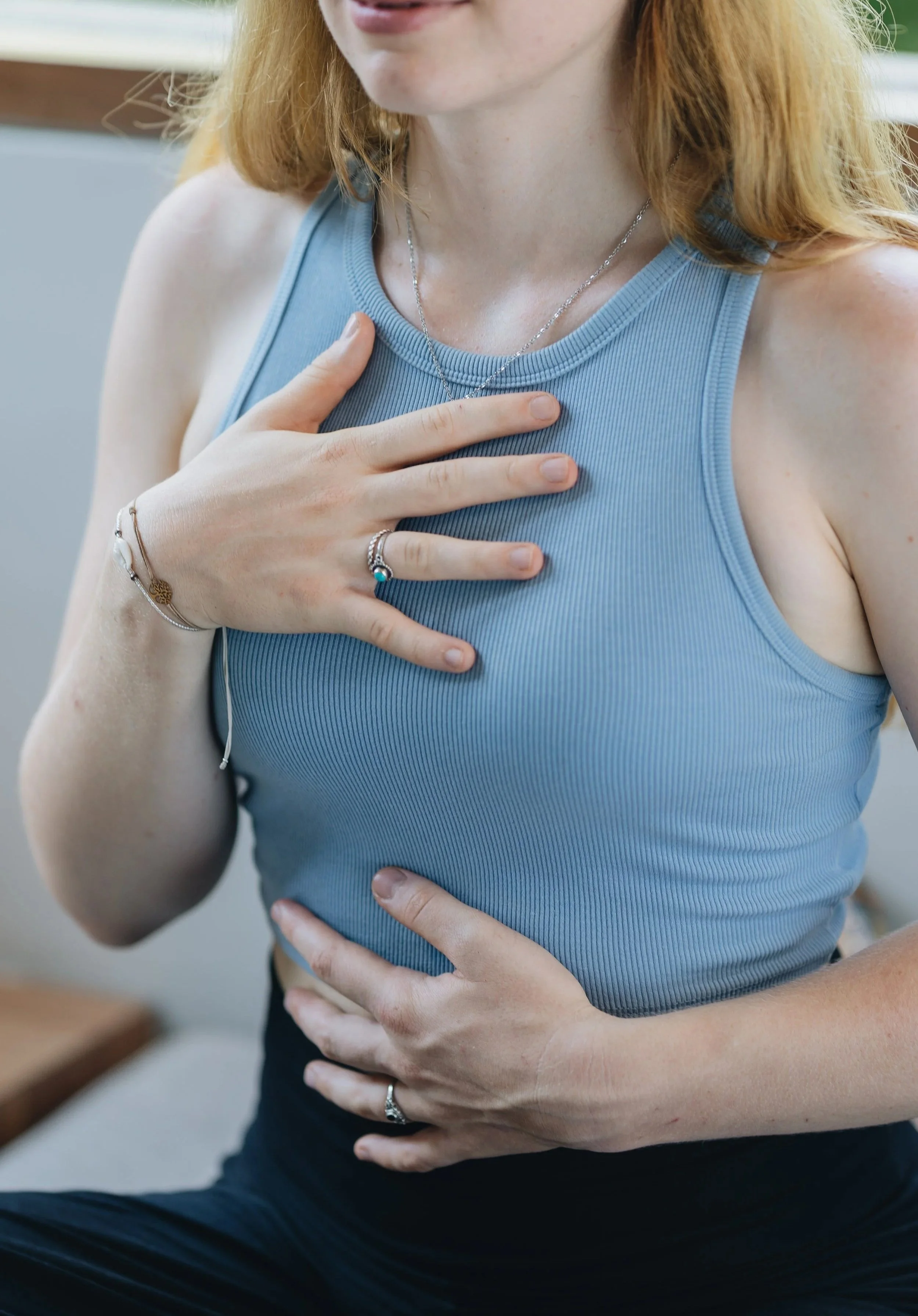 A woman with red hair wearing a blue tank top, holding her chest and stomach, with her left hand resting on her stomach and right hand on her chest. She is wearing rings and a necklace.