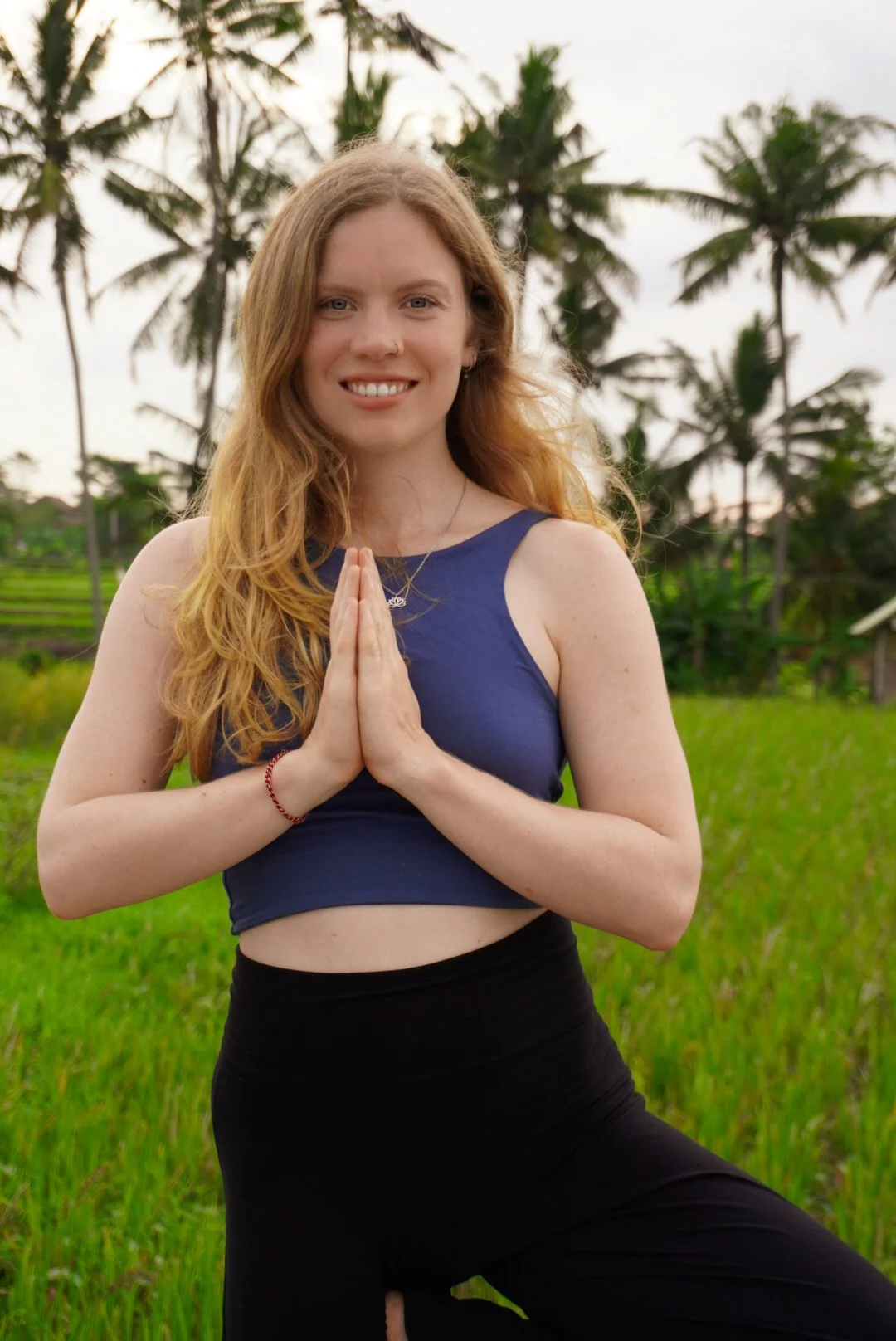 A woman practicing yoga in a lush outdoor setting with palm trees in the background, wearing a blue crop top and black pants, with hands in prayer position.