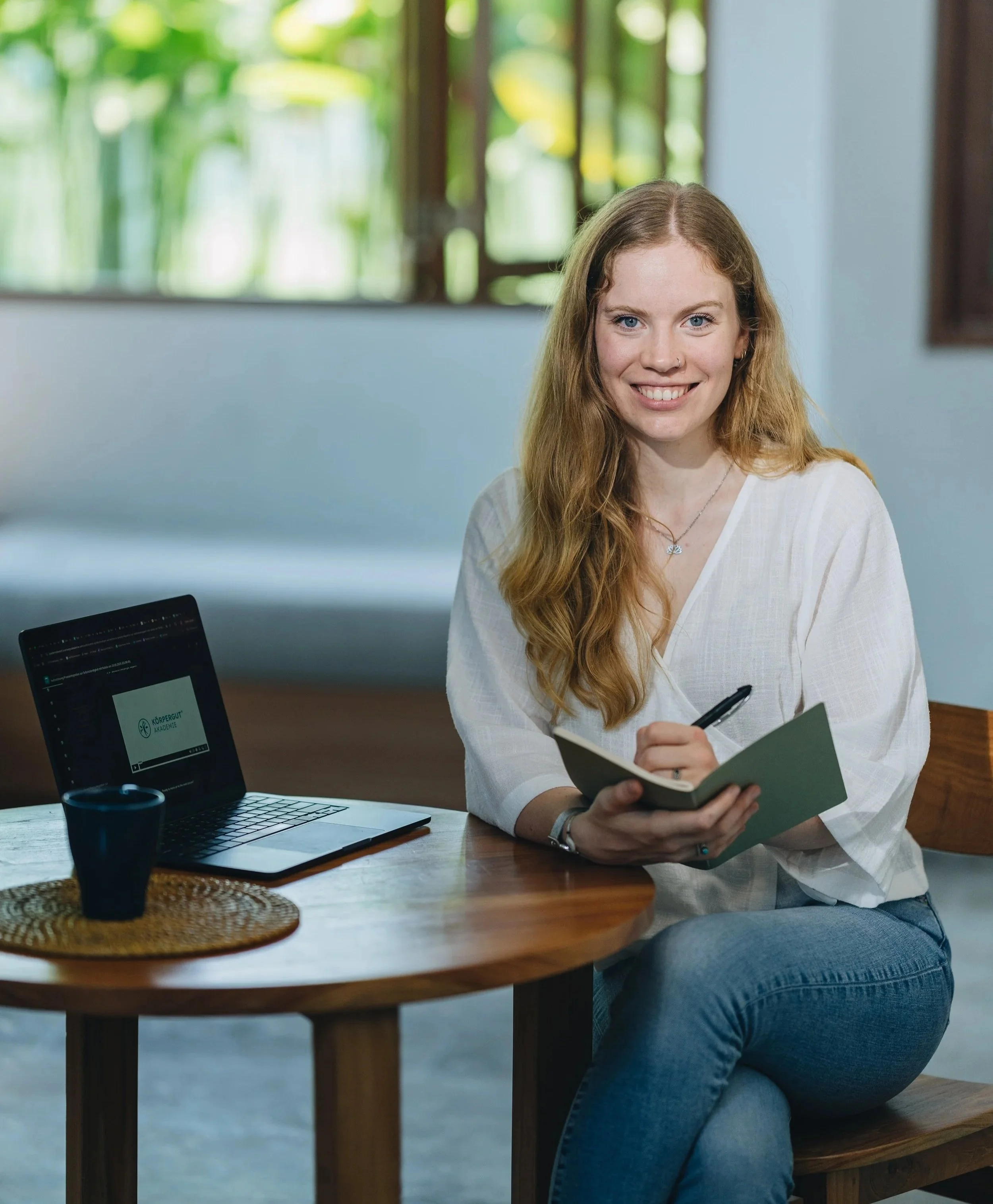 A young woman with red hair and a white blouse sitting at a wooden table, smiling, with an open notebook and pen in her hands. A laptop and a black mug are also on the table, in a bright room with windows showing greenery outside.