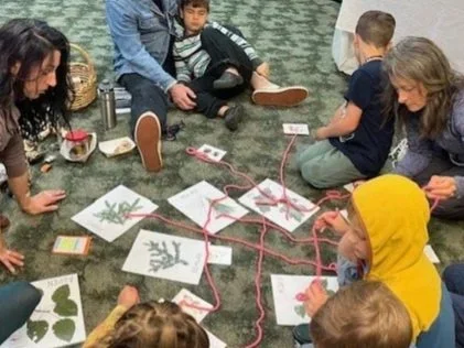 A young girl kneeling on the ground in a garden, planting seeds in a small pot, with gardening tools, plant soil, and gardening supplies around her.