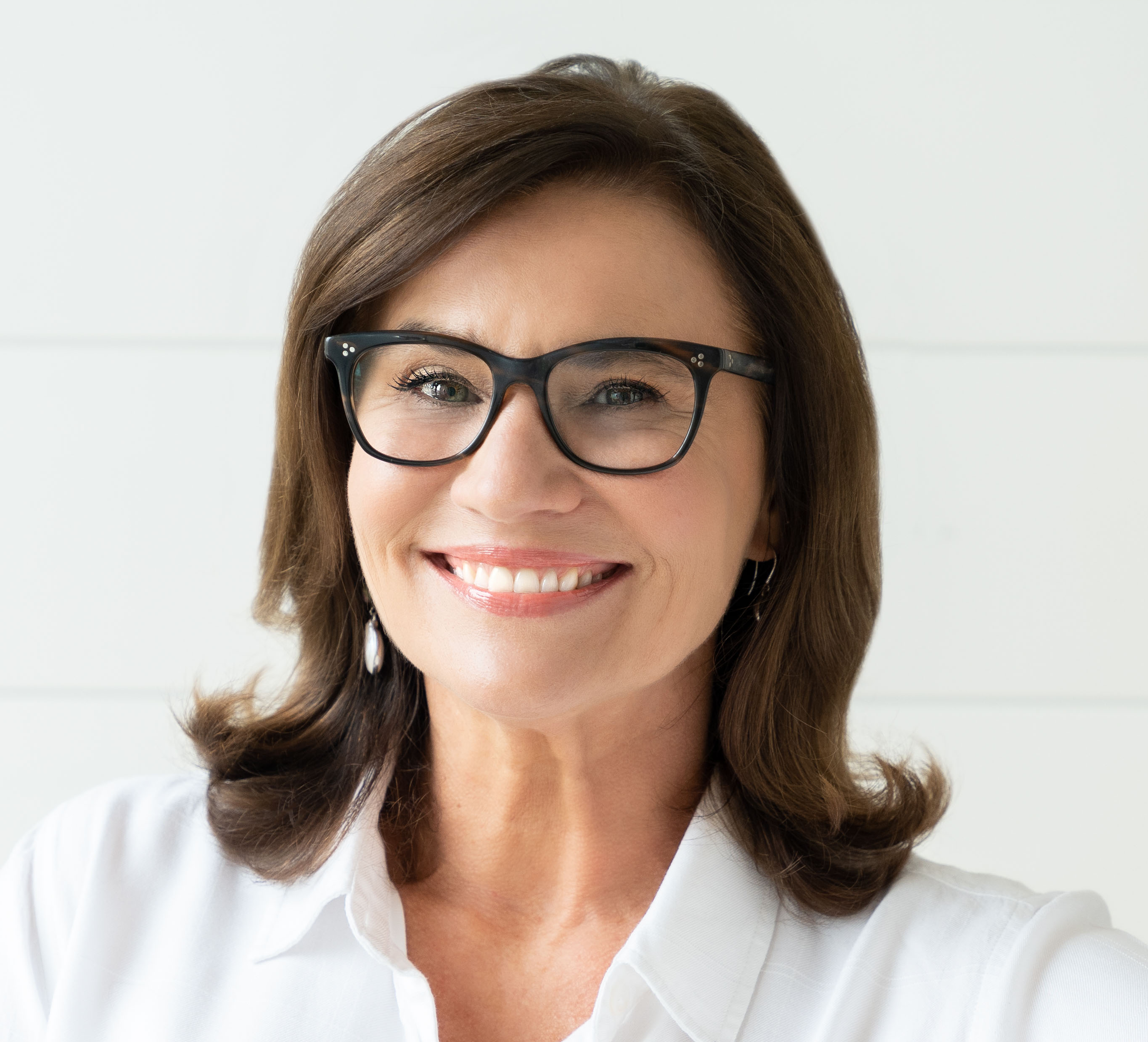A woman with shoulder-length brown hair, glasses, and earrings smiling at the camera, wearing a white blouse, with a plain white background.