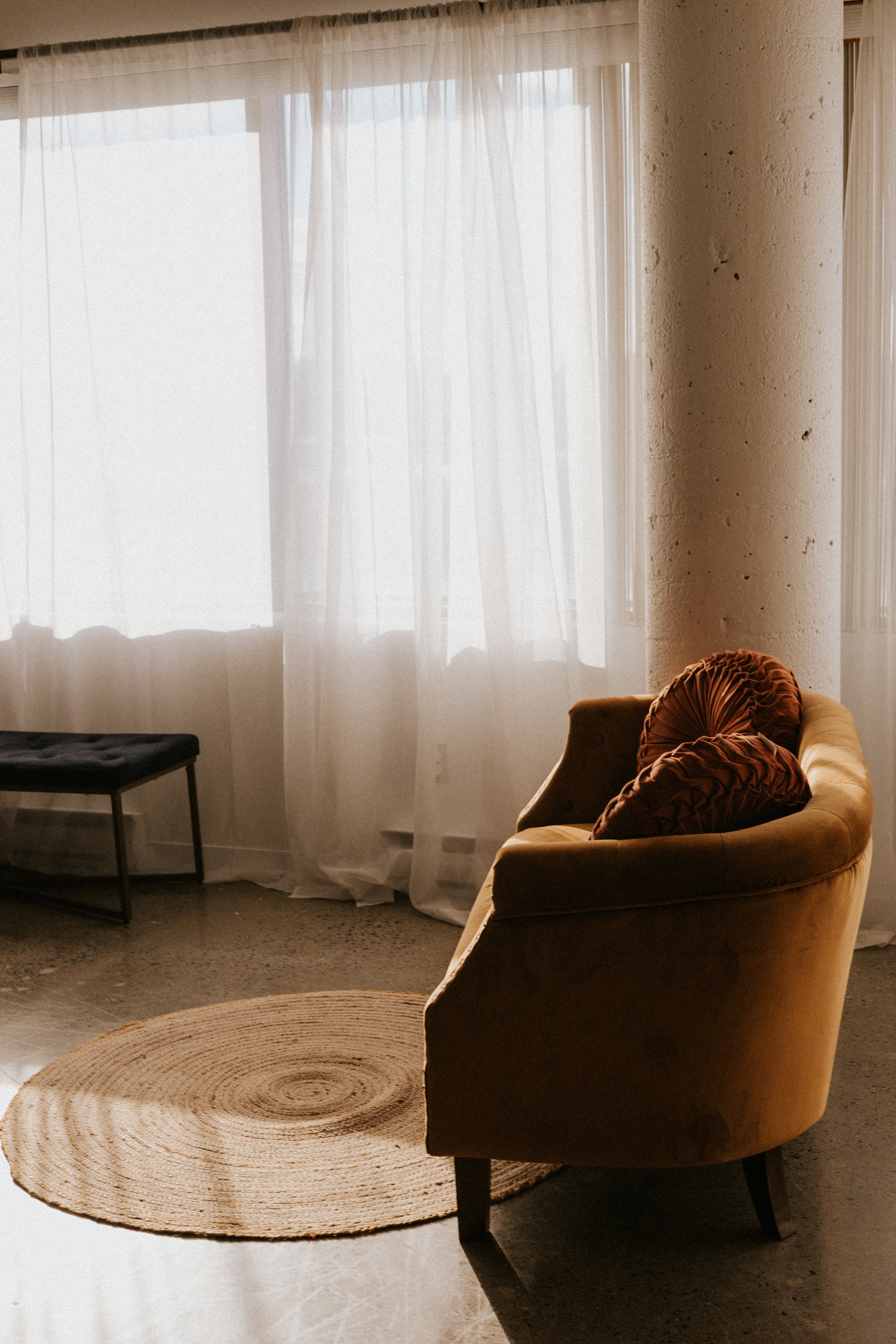 A cozy corner with a beige velvet armchair with two rust-colored pillows, a circular woven rug, sheer curtains, and a black bench by the window, illuminated by natural sunlight.