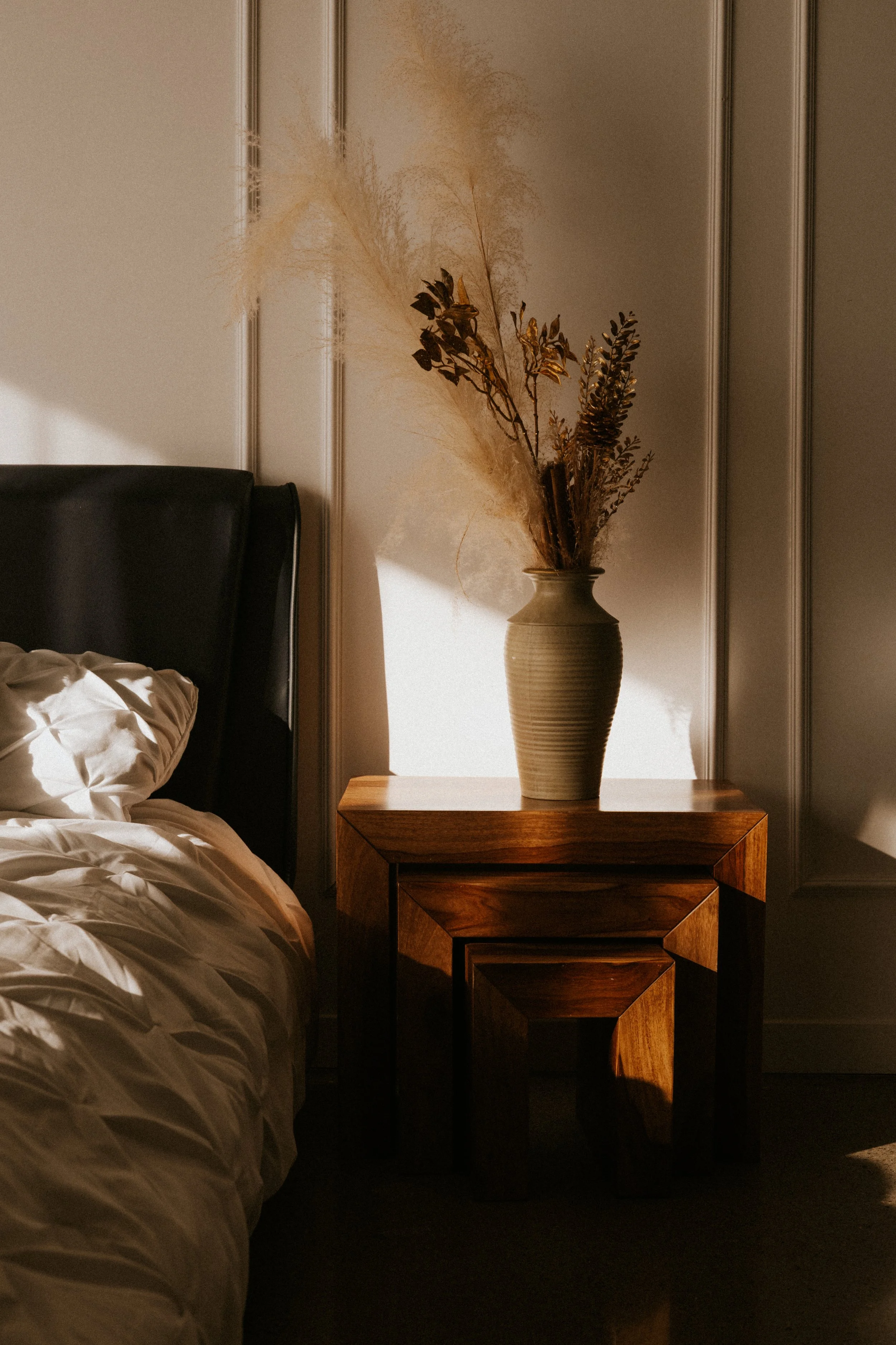 A bedroom scene with a black headboard and white bedding on the left, a wooden bedside table on the right, and a beige vase with dried plants on top. Sunlight casts shadows on the wall.