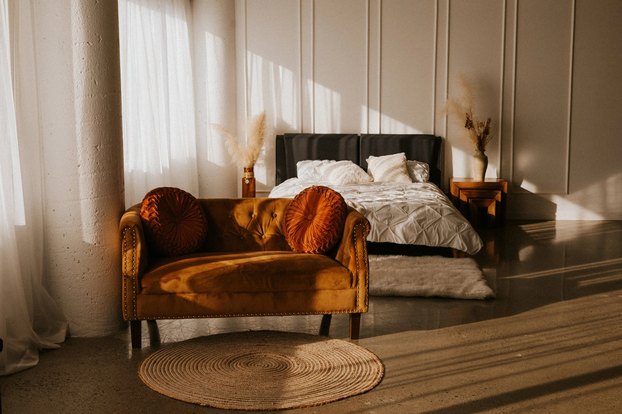 Sunlit bedroom with a bed against the wall, a vintage velvet couch with two round pillows in front, and a small wooden side table with a vase of dried plants on each side of the bed.
