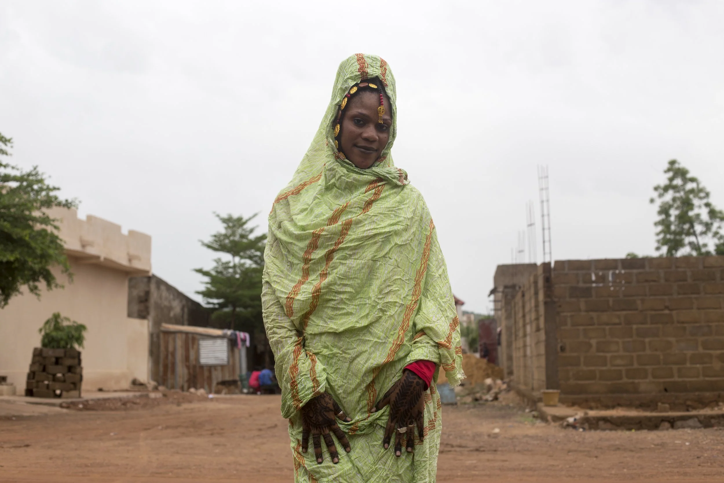A woman in a green traditional dress with patterns, wearing jewelry and henna on her hands, standing outdoors in a rural area with simple buildings and trees in the background.