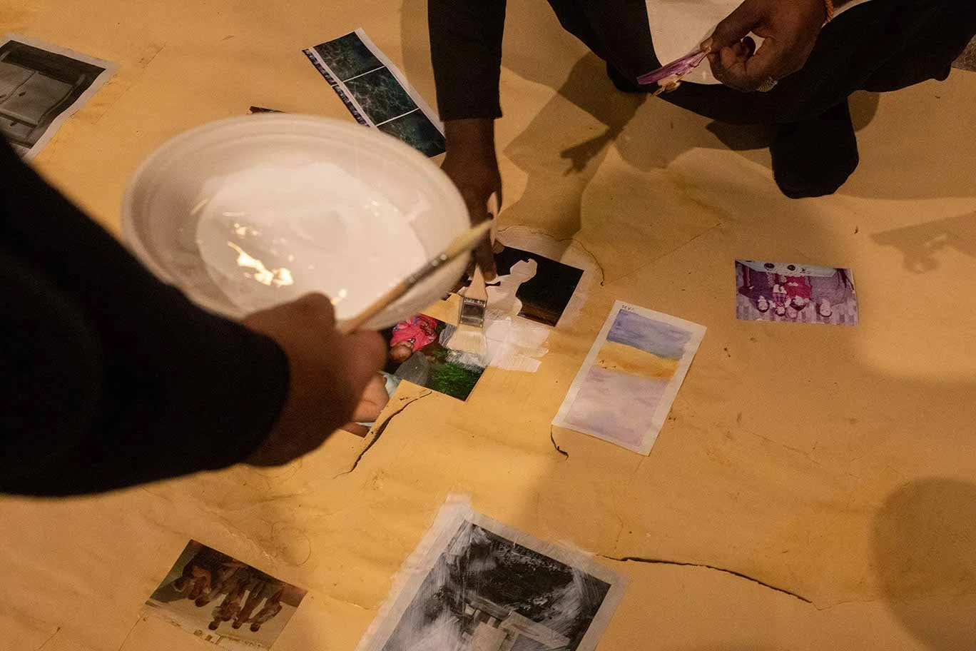 People looking at photographs spread out on a table, one person holding a bowl with a brush in it.