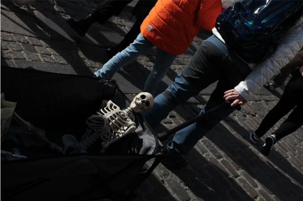 A small skeleton toy is placed on a stroller, surrounded by people walking outdoors on a cobblestone street.