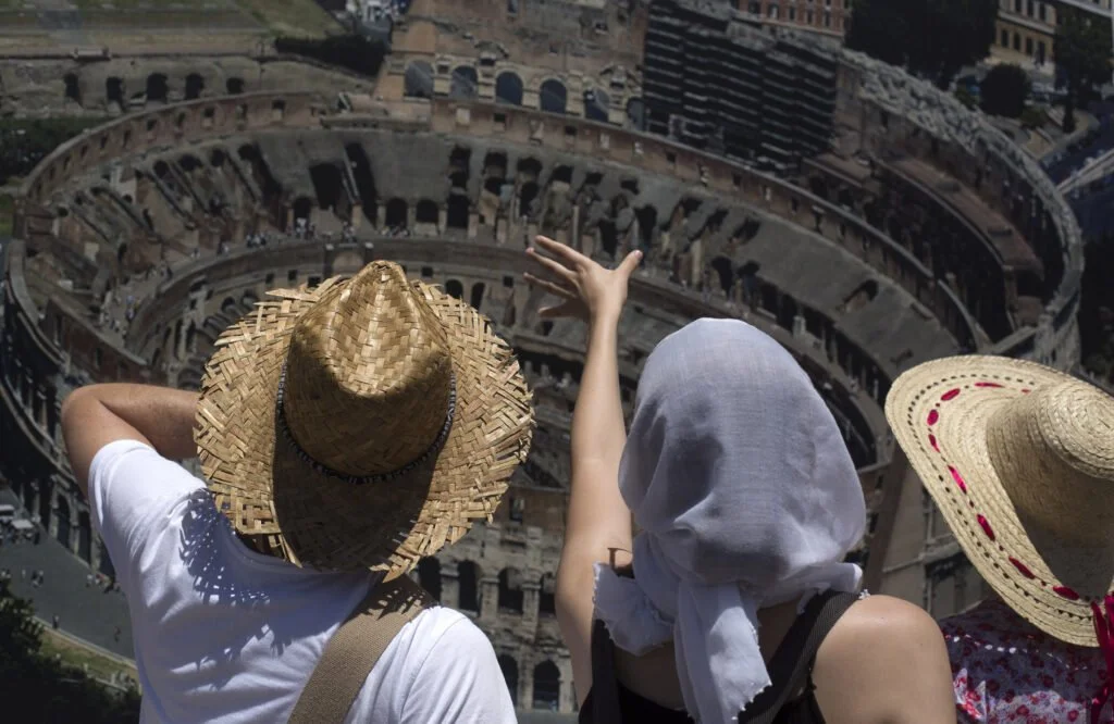 Three tourists wearing wide-brimmed straw hats and summer clothing overlook the Colosseum in Rome, Italy.