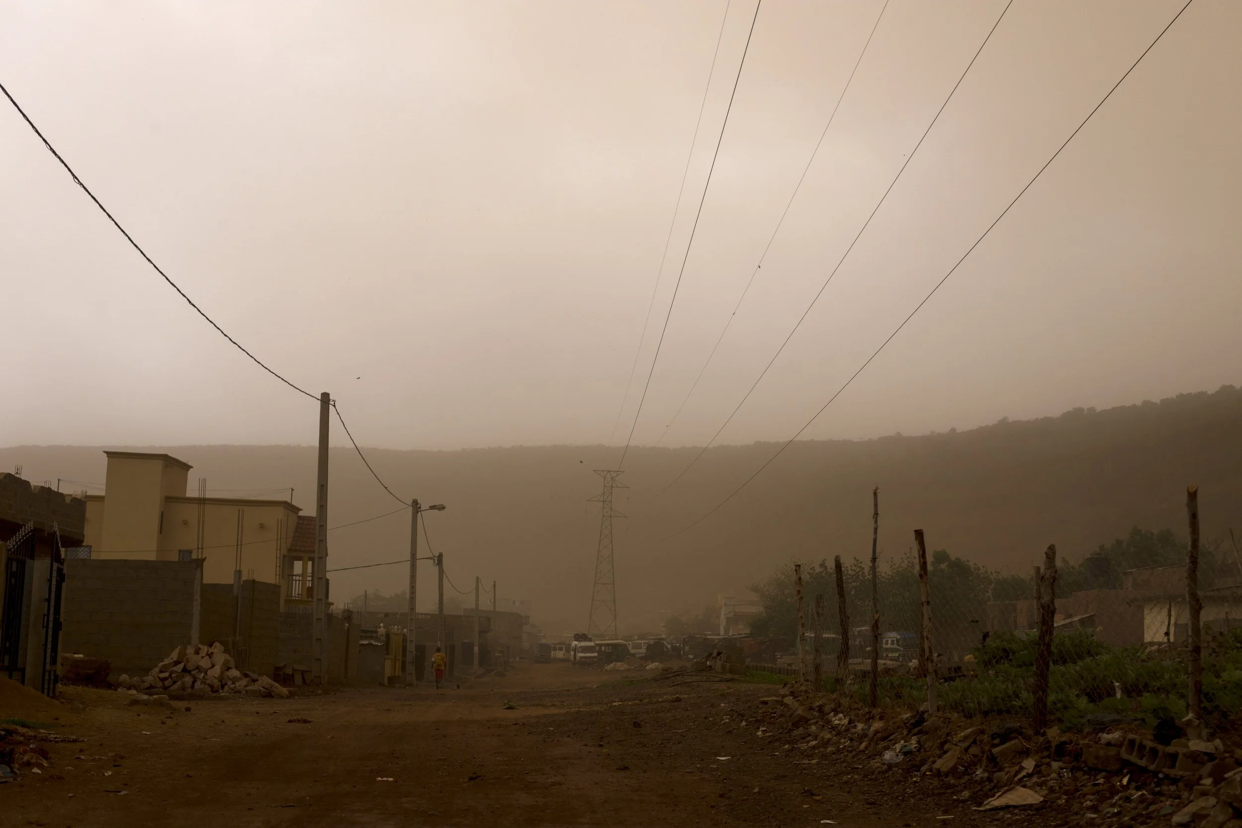 A dusty, unpaved road in a rural area with utility poles, power lines, and some houses along the sides. A hazy, overcast sky covers the landscape.