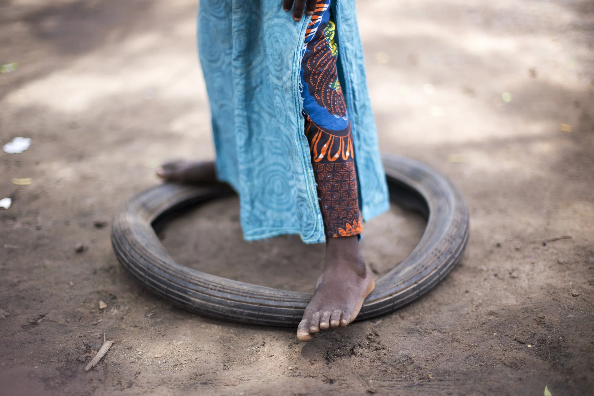 A person standing barefoot on the ground, with one foot on a tire, wearing colorful patterned clothing and a blue towel or cloth draped over their leg.
