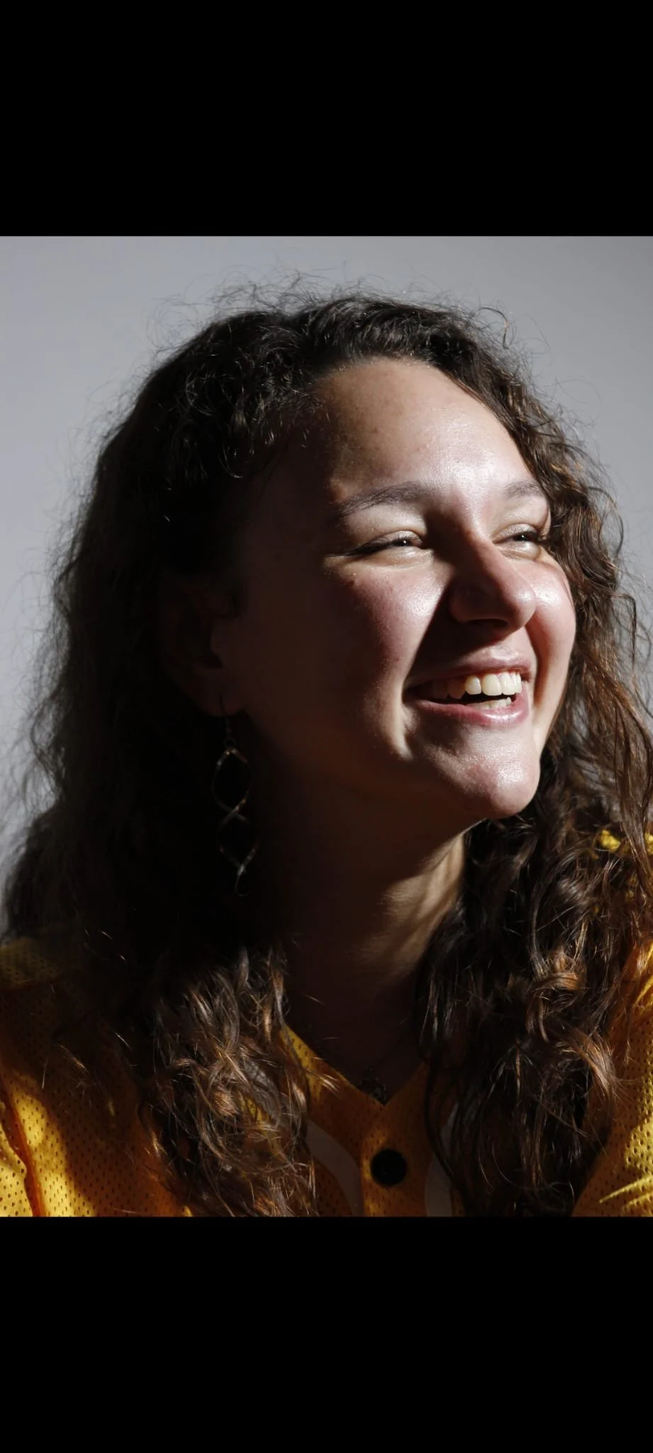 A woman with curly hair smiling and wearing a yellow sports jersey.